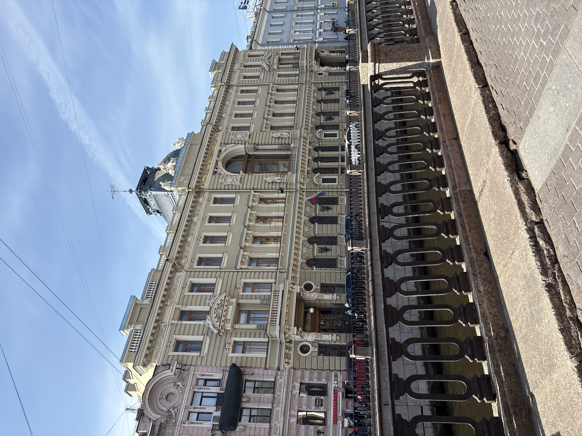 An upward-angle, exterior shot shows the headquarters of the "First Society of Mutual Credit" in Saint Petersburg, Russia. The building, primarily tan, features multiple floors and ornate architectural details. The foreground includes a dark metal fence, a sidewalk, and several parked vehicles. Above the building, a clear blue sky hosts faint clouds and power lines.