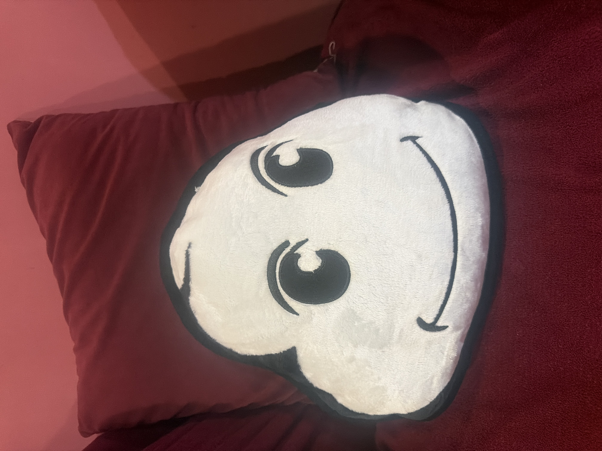 A white, cloud-shaped pillow with a black outline and a smiling face is positioned on top of a red pillow. The pillow is on a red surface. The location is unknown.