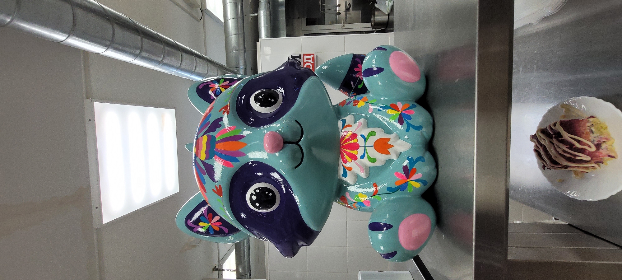 A decorated, cartoonish statue of a panda bear lies on its side. The panda is painted light blue with dark purple around the eyes and head. Its ears and feet are light blue with pink accents. The body features colorful floral designs. It is laying on a stainless steel counter next to a white plate of food with a drizzled sauce. Above, there is a row of fluorescent lights and metal ductwork. There is a sign in the background, partially obscured by the panda. The location is Unknown, Unknown.