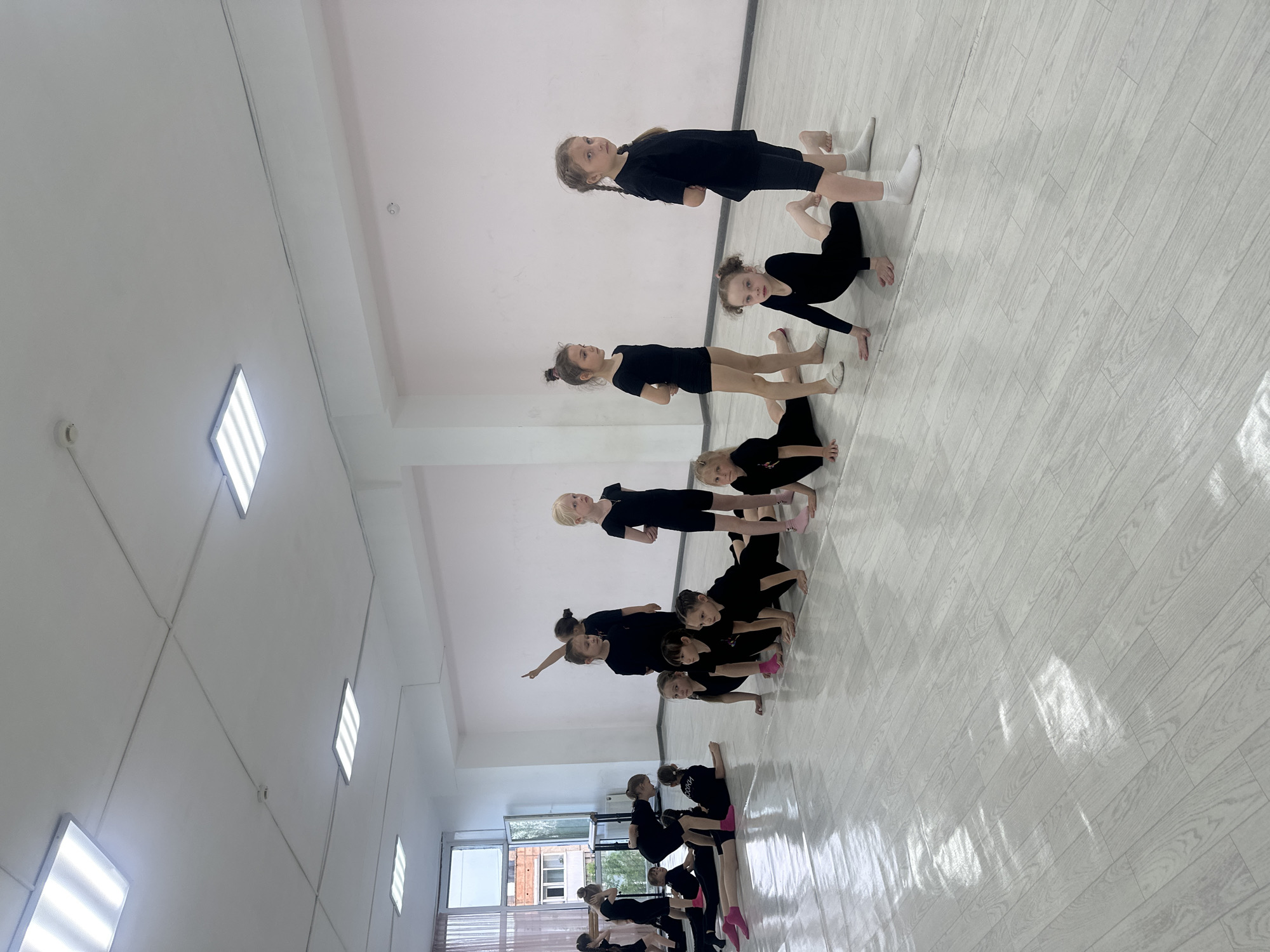 The image shows a group of young girls in black athletic wear practicing gymnastics or dance in what appears to be a dance studio or gymnasium. The girls are arranged in a line performing a coordinated pose, with some girls supporting others in elevated positions. The studio has white walls and a light-colored, wood-patterned floor. The lighting is bright, likely from overhead fluorescent lights. A window is visible in the background, showing a view of trees and buildings outside. Based on the context provided, the location is Votkinsk, Russia.