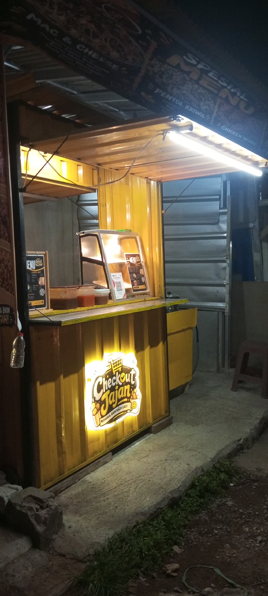 The image depicts a food stall in Setu, Indonesia, at night. The stall is constructed from yellow corrugated metal and has a brightly lit "Checkout Jajan" logo on its front. Above the stall, a sign advertises "MAC & CHEESE" and "SPESIAL MENU" with listed items like "PENTOL KRIWIL" and "CHICKEN WINGS." Inside the stall, menus are displayed on a counter, along with containers of sauces. The scene is illuminated by overhead fluorescent lighting, casting a warm glow on the yellow structure. The ground in front of the stall is concrete, with some patches of grass and debris. A small red stool is visible to the right of the stall. The overall impression is of a small, local food vendor operating after dark.