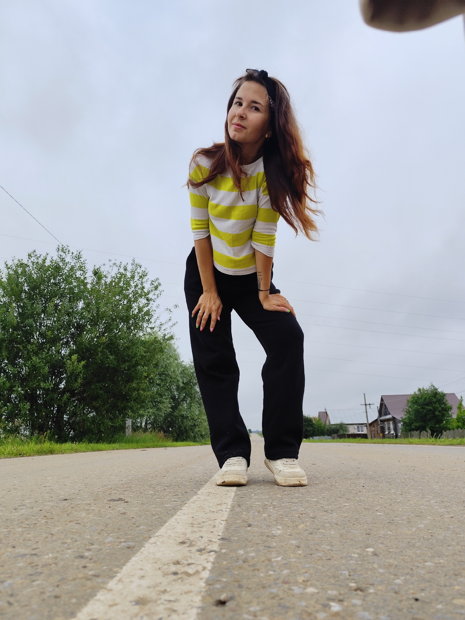 A woman with long, brown, wavy hair, wearing a yellow and white striped long-sleeved shirt and black pants, is standing on a paved road. She is leaning forward with her hands on her knees and looking at the camera. The road has a white line painted down the center. In the background, there are trees on the left and houses on the right. The sky is overcast and gray. The location appears to be the city of Fokino, Russia.