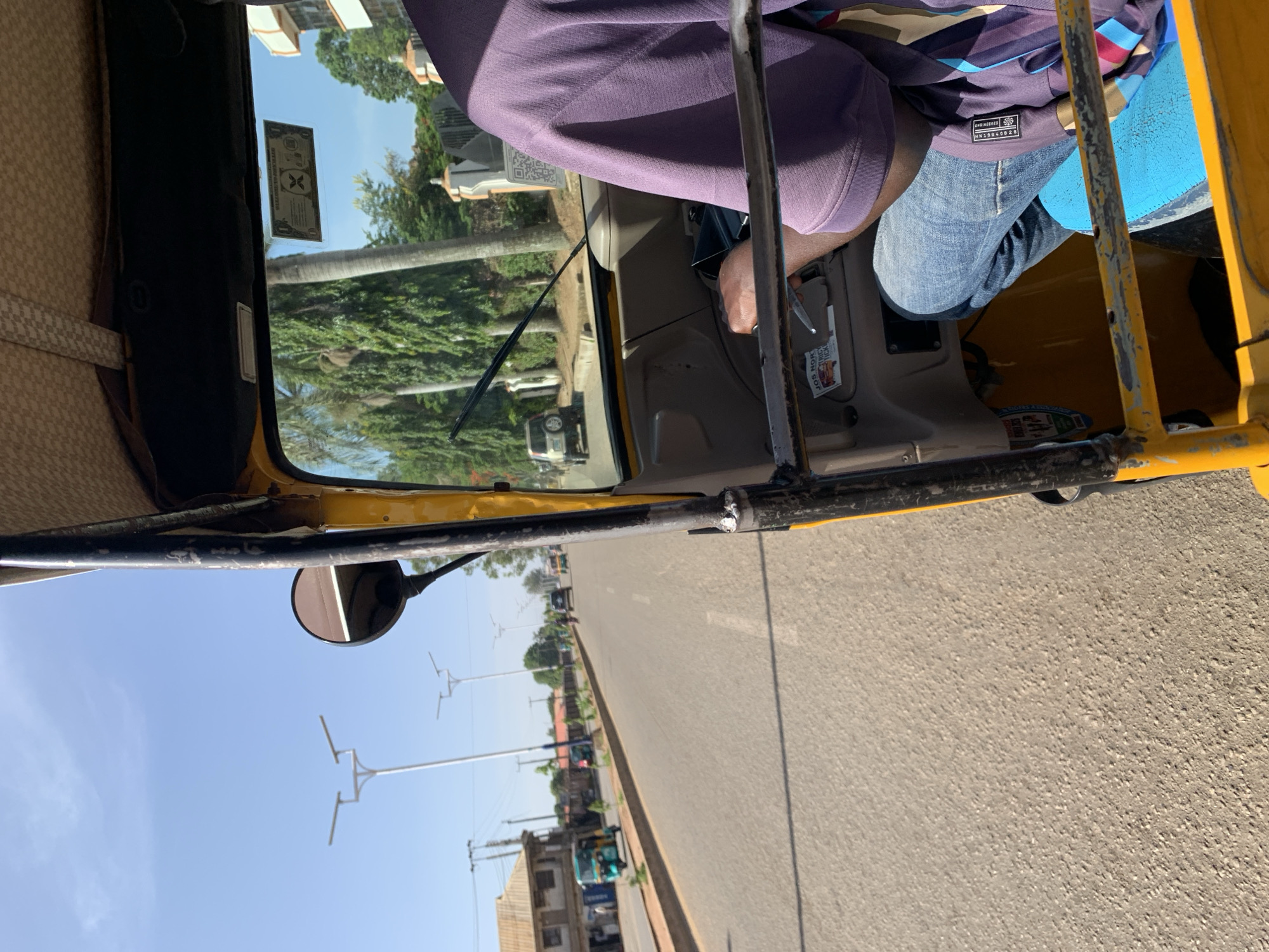 The image depicts a first-person perspective from inside an auto-rickshaw (often called a 'keke napep' in Nigeria) in Rafin Par, Nigeria, during daylight hours. The camera is positioned to show both the vehicle's interior and the street scene ahead.
A person, likely the driver or a front passenger, is prominently featured from the mid-torso down. They are wearing a purple short-sleeved top with a patterned shoulder section and blue jeans. A clothing label on their top reads "ENGINEERED". Their right arm and hand are visible near the vehicle's controls. A small sticker attached inside the vehicle, near the driver's area, has partially legible text including "JOS NON STOP" and "BEST QUALITY".
The interior of the auto-rickshaw shows part of the dashboard, a side mirror, and a sturdy yellow frame structure. On the windshield, a rectangular official-looking document or sticker is displayed, with "NIGERIA..." faintly visible at the top and a prominent 'X' shaped emblem below.
Beyond the vehicle, a paved asphalt road extends into the distance under a clear, bright blue sky, indicating sunny weather. The roadside is lined with green trees, some sparse buildings, and modern streetlights featuring multiple horizontal arms. Other distant vehicles can be seen further down the road, suggesting a populated area. The overall scene conveys a typical street view from a moving or stationary vehicle in an urban or semi-urban setting in Nigeria.