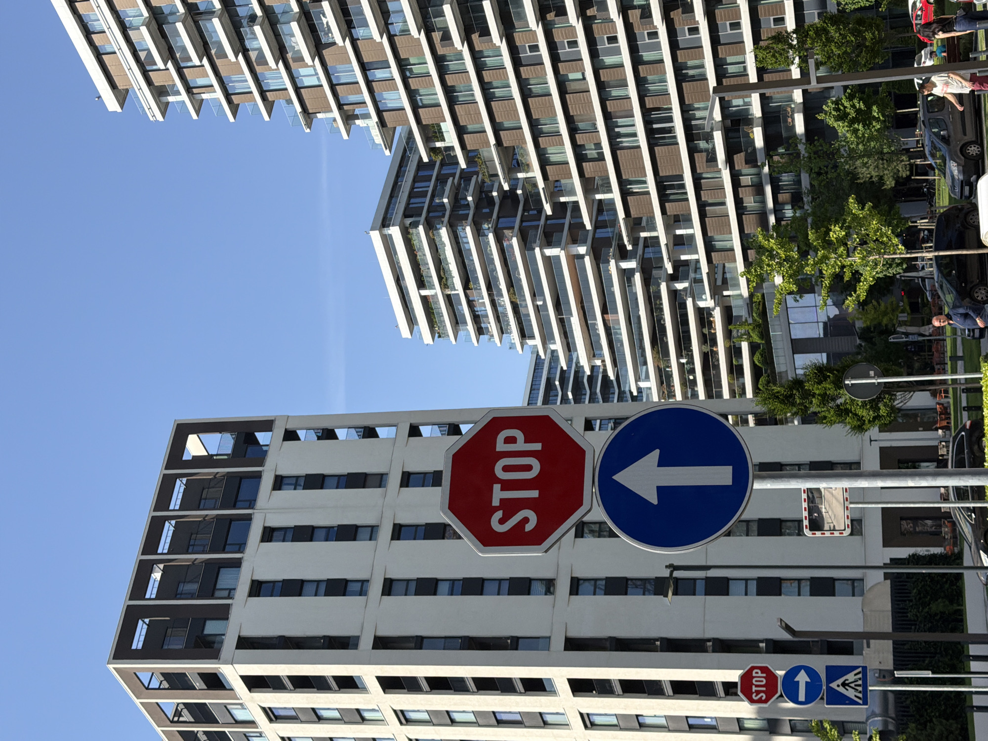The image captures a street scene in Belgrade, Serbia, focusing on architectural elements and street signs.
In the foreground, there are two prominent buildings. One has a design of protruding balconies, while the other features a grid-like structure with alternating dark and light horizontal bands. A pair of traffic signs are suspended in front of the lower building. One is a red octagonal stop sign, and the other is a blue circular sign with a white arrow pointing upwards.
The backdrop features additional buildings, trees, parked cars, and pedestrians. The sky is a clear blue. The buildings' architecture appears modern, with rows of windows and geometric designs. The image is rotated.