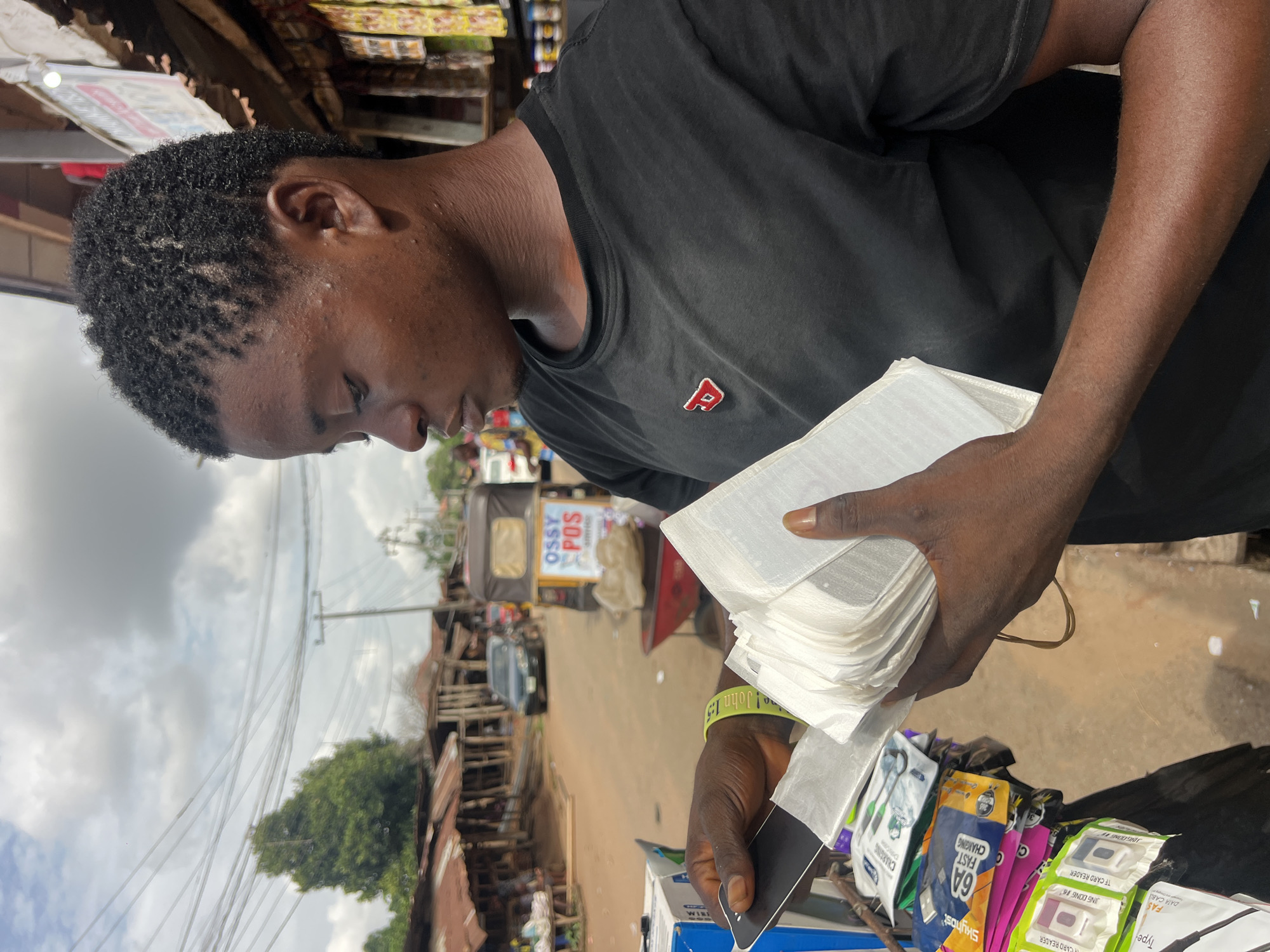 A man stands in what appears to be an urban street or marketplace in the city of Unknown, Unknown. He is holding a stack of what seem to be phone screens or protectors, possibly for sale, given the assortment of tech accessories visible on a table or cart in front of him. The man is dark-skinned, wearing a black T-shirt with a small red emblem on the chest. He has short, dark hair and appears to be focused on the items in his hands. In the background, there's a street scene with various buildings, vehicles including a tricycle, and signs indicating commercial activity. Power lines stretch across the sky.