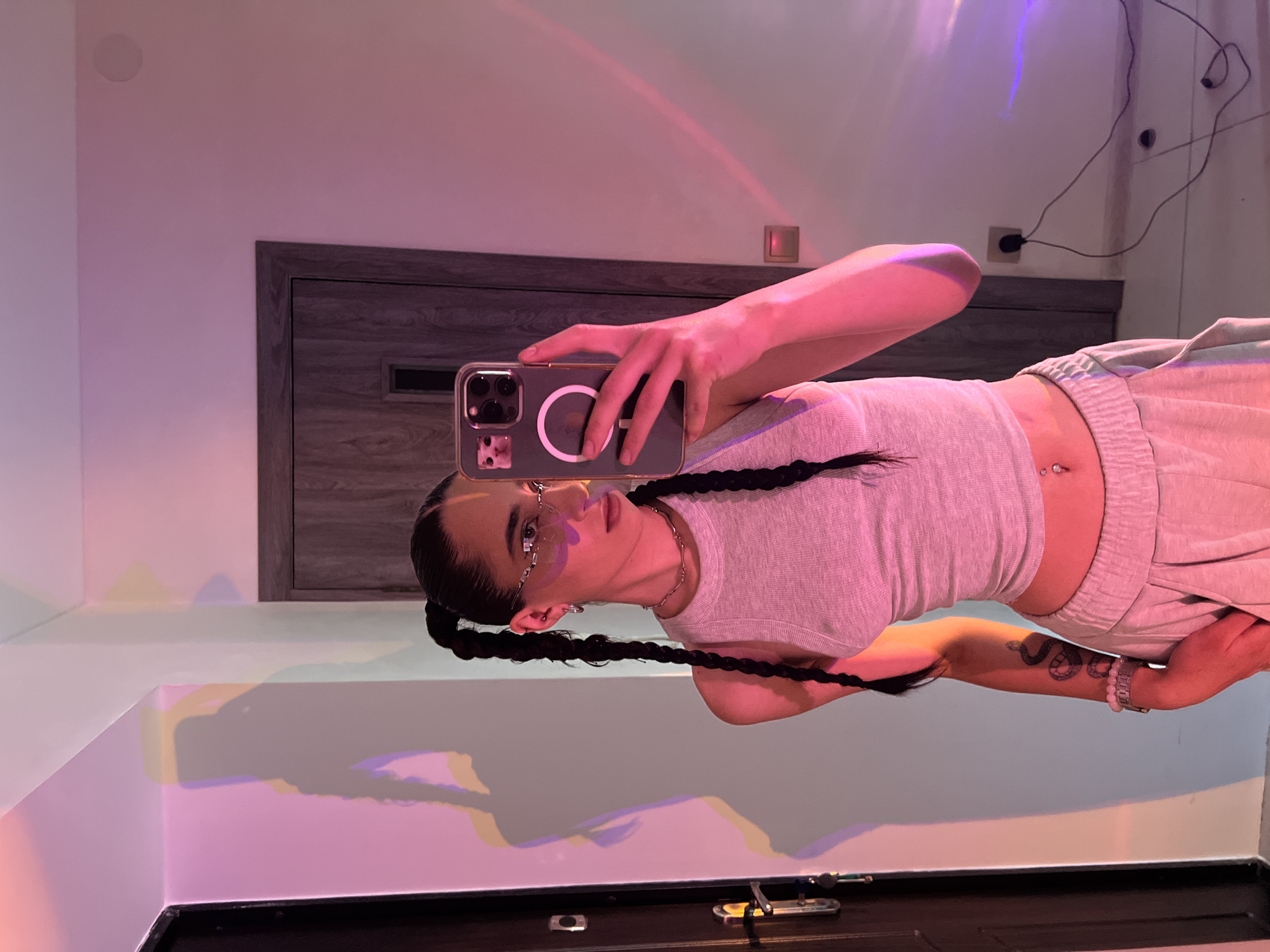 A young woman with dark hair braided into two pigtails is taking a selfie in a room. She is wearing a light-colored sleeveless top, light-colored sweatpants, and distinctive silver glasses. Her left arm has a tattoo of a snake. A phone with a cat sticker is held in her right hand, capturing the reflection. The room has a door with a dark wood finish and a light-colored wall. Colorful lights are projected onto the wall, creating shadows. The scene appears to be an indoor residential setting, possibly an apartment. The presence of a plug and cables suggests a power outlet is nearby. There is no discernible information in the image that would indicate the location is Ternopil, Ukraine.
