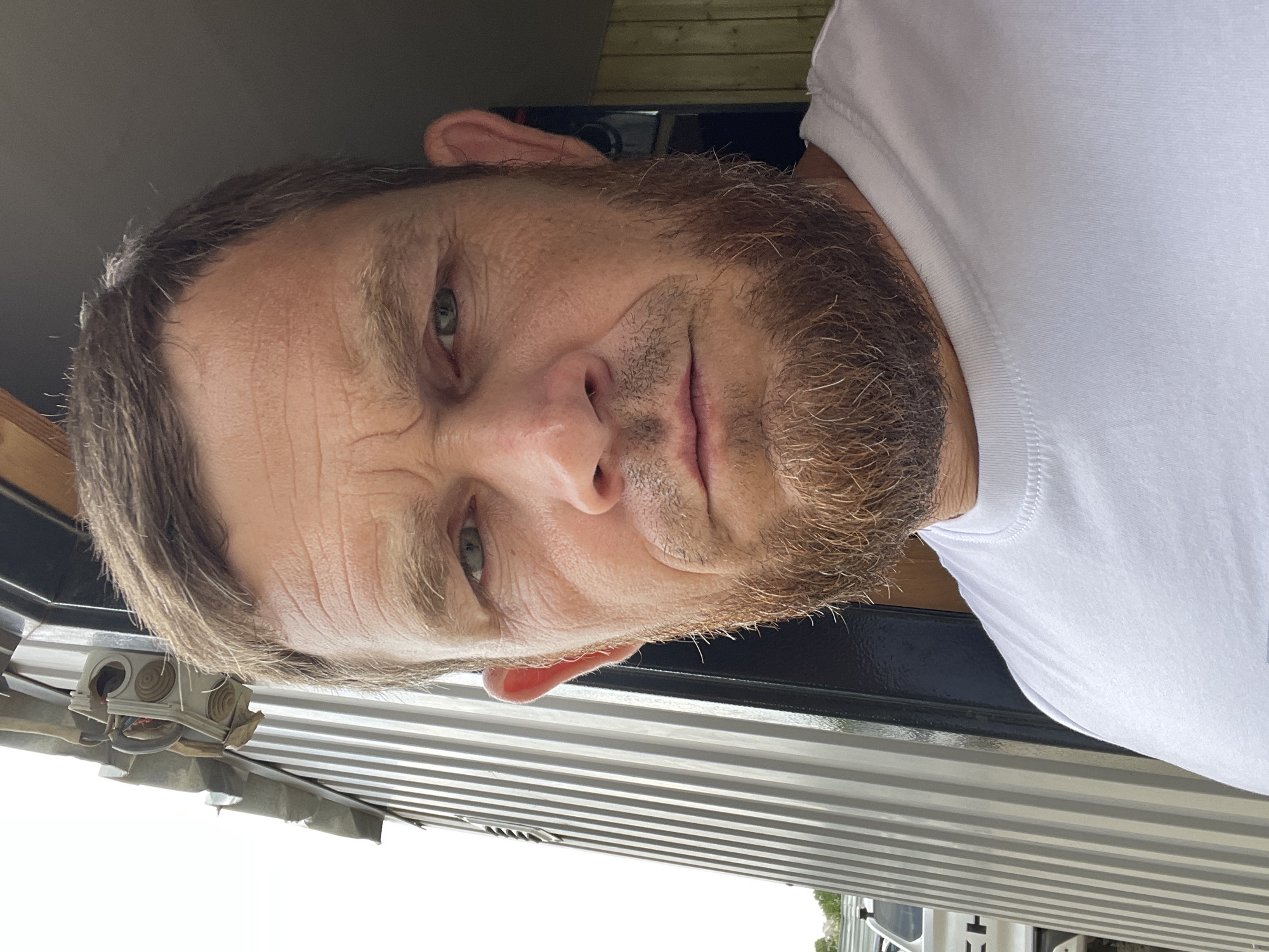 A close-up, head-and-shoulders shot of a man with a beard and a white t-shirt is presented. The man has fair skin, light brown hair, and greenish-blue eyes. His facial expression appears neutral or slightly serious. He has a noticeable beard and mustache, and his forehead shows some wrinkles. The image is oriented vertically, with the man's head at the top and his body extending downwards.
The setting seems to be outdoors, possibly in a backyard or a workshop area. In the background, a dark-colored surface, possibly a wall or a vehicle, is visible. To the left and bottom of the frame, there are horizontal striped surfaces, suggesting corrugated metal or siding. A small electrical junction box with wires is also visible near the bottom left, attached to the corrugated surface.
The lighting suggests it is daytime, likely with overcast or diffused sunlight, as there are no harsh shadows. The overall mood is candid and informal. No text is visible in the image. The location context provided is Krasnodar, Russia, but there are no specific visual cues within the image to confirm this.