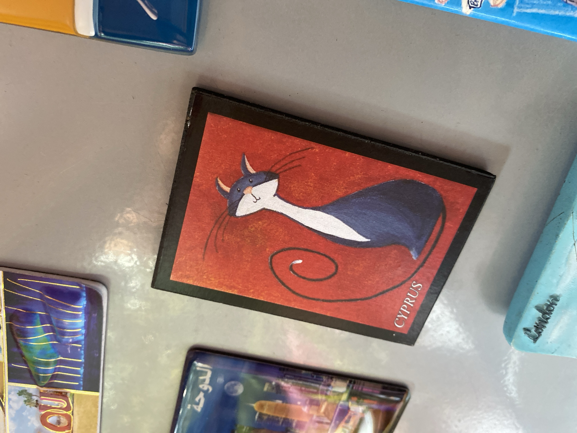 The image shows a close-up of a collection of refrigerator magnets. In the foreground, a black-bordered magnet features a stylized illustration of a dark blue cat with white markings against a vibrant red background. The text "CYPRUS" is visible in white at the bottom of this magnet. To the left, another magnet displays a cityscape with tall buildings and Arabic text that reads "الدوحة" (Doha). Above this, a portion of another magnet shows colorful, striped structures. Partially visible on the right is a magnet with a blue and yellow design, and further down, a light blue magnet with the word "London" inscribed. The background is a plain, grey, metallic surface, consistent with a refrigerator door. There are no people or animals present in the scene, nor any indication of the city of Wrocław, Poland.