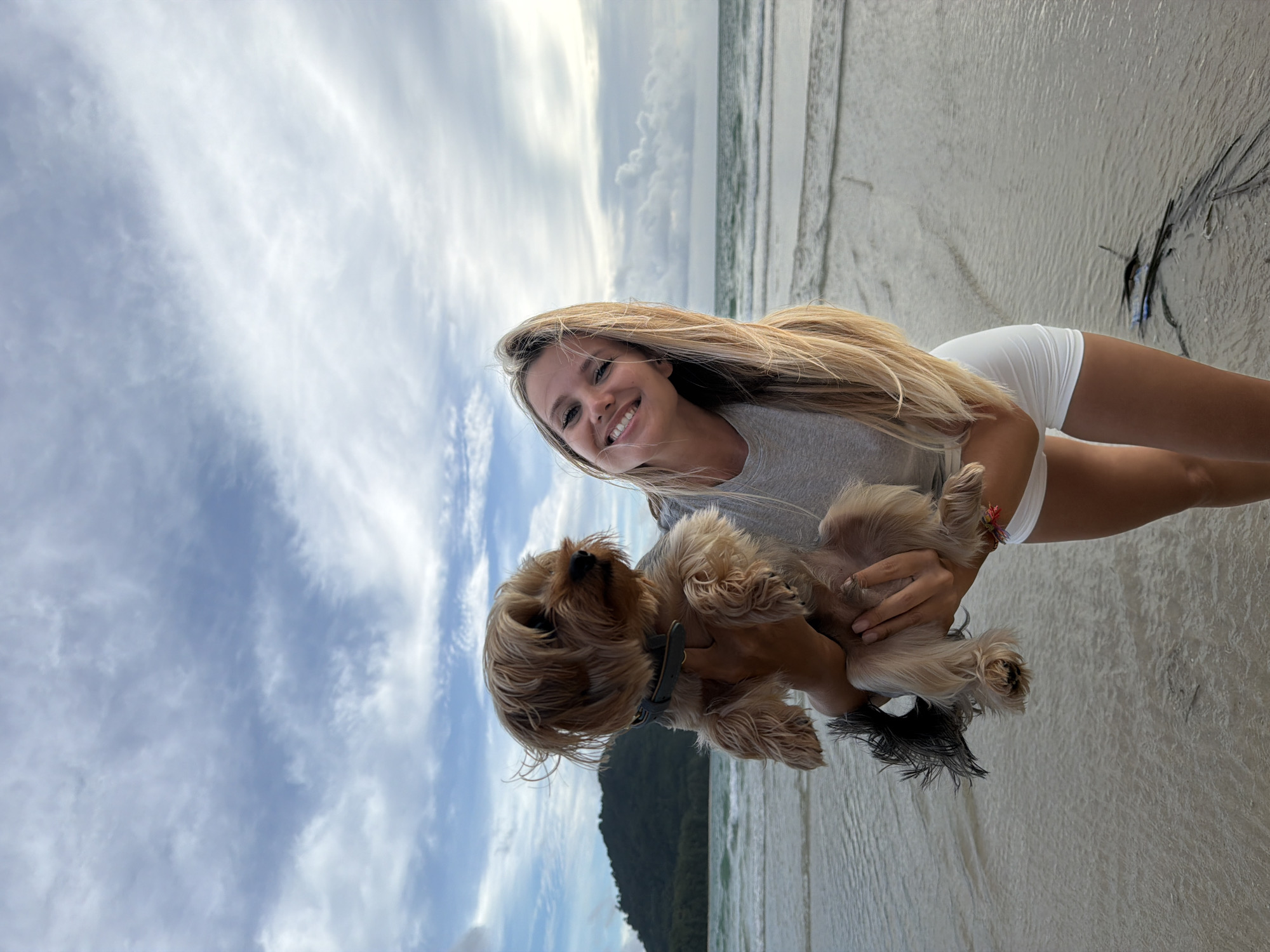 A young woman with blonde hair and a bright smile is holding a small dog on a beach in Sakhu, Thailand. The woman is wearing a grey t-shirt and white shorts. The dog is a light brown Yorkshire Terrier with a grey collar. The background shows the ocean with gentle waves and a lush green island in the distance under a cloudy sky. The sand is wet from the receding tide, and there are some dried seaweed and debris scattered on the beach. The overall scene is one of happiness and relaxation in a tropical setting.