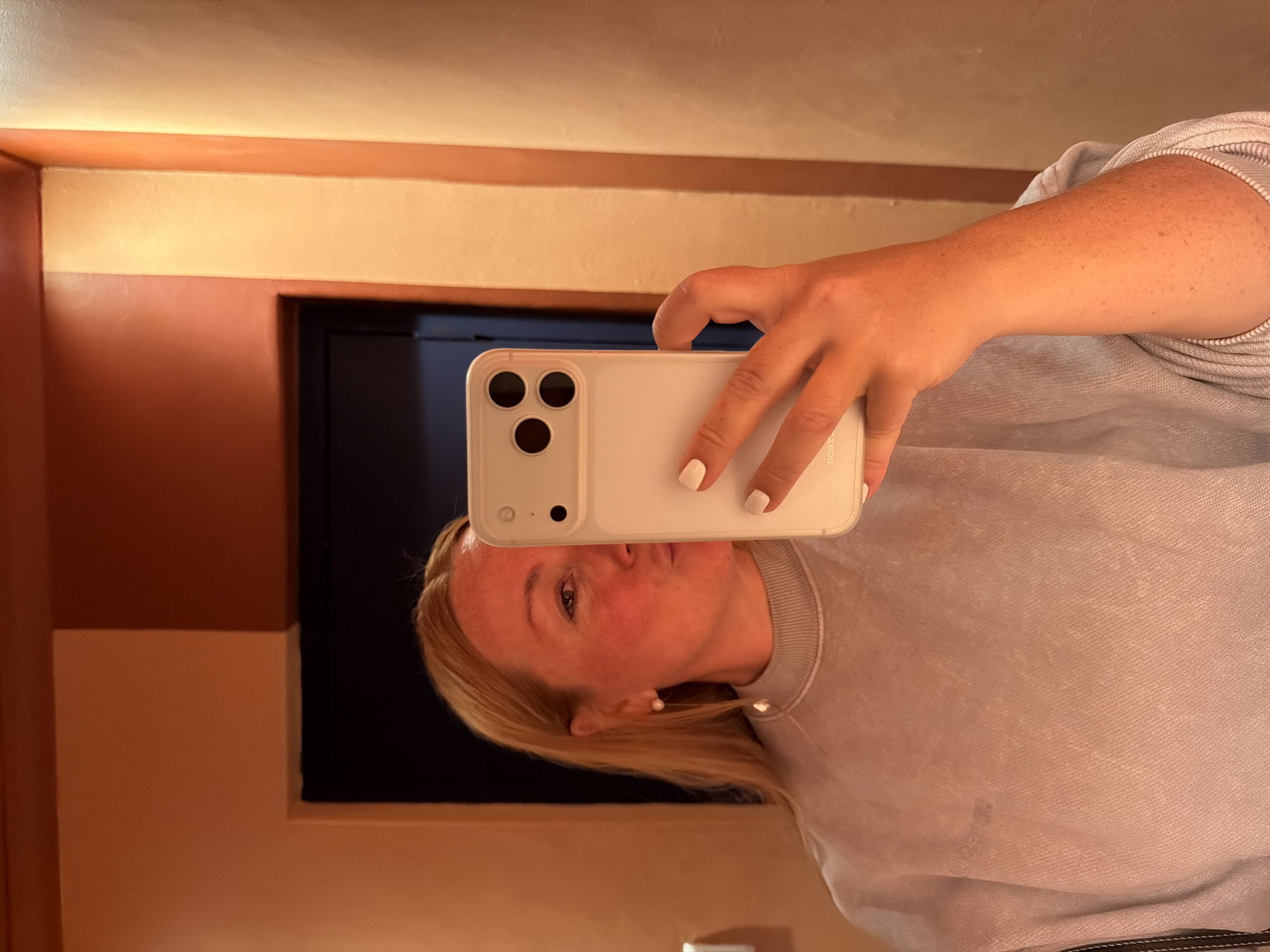 A woman with blonde hair is taking a selfie with her white smartphone. She is wearing a light gray sweatshirt and a pearl earring. The background features a wall with alternating bands of beige and brown paint. A dark rectangle, likely a mirror or a window, is visible behind her. There is no clear indication of the city of Cape Town, South Africa in this image.