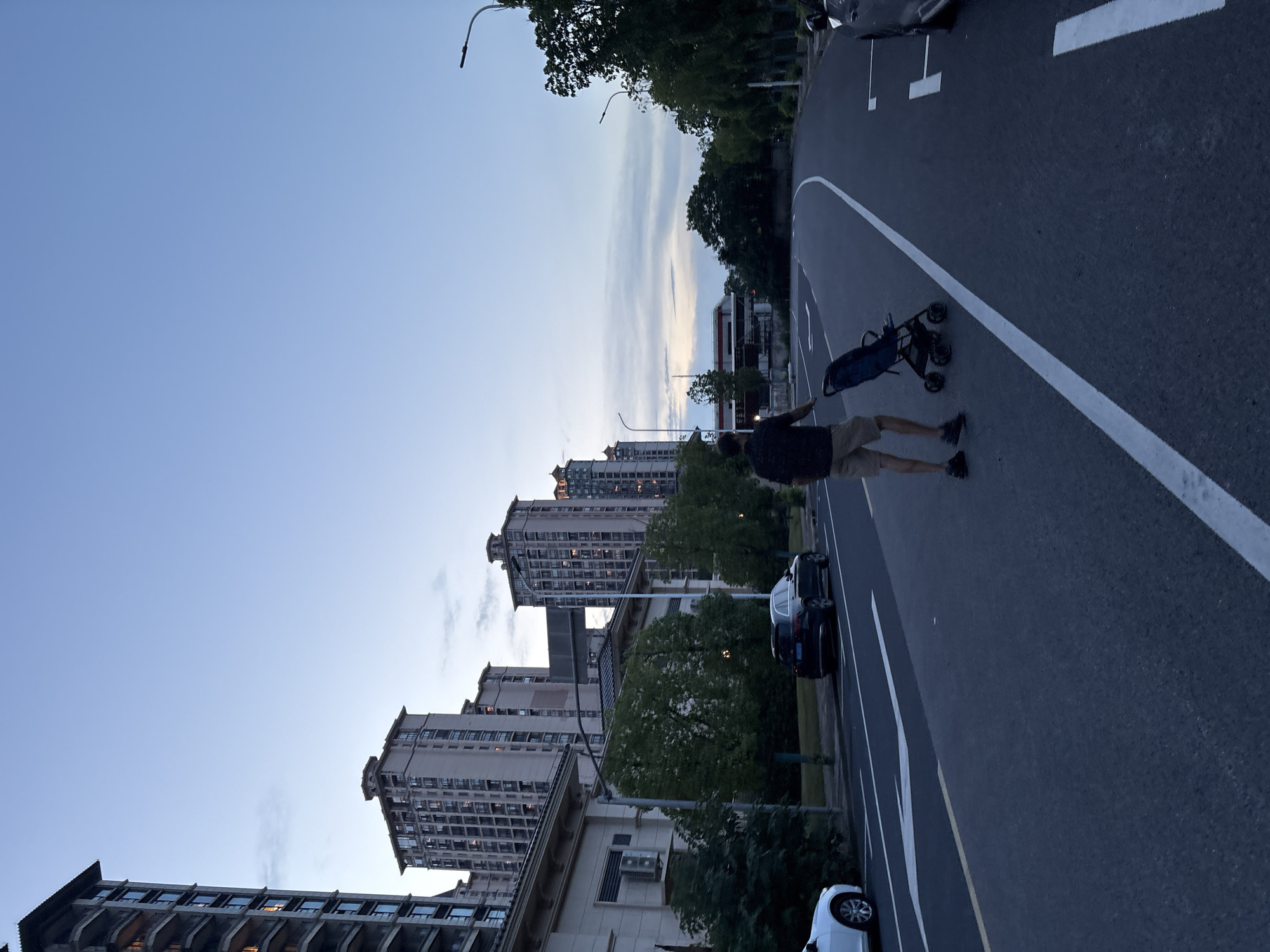 In Minhang District, China, a man is pushing a stroller on a road lined with trees and apartment buildings. The sky is a pale blue with some scattered clouds, suggesting it is either early morning or late evening. The road has white lane markings and a yellow line separating traffic lanes. Cars are parked along the side of the road, and the apartment buildings appear to be modern high-rises with many windows. The scene has a quiet, residential feel.