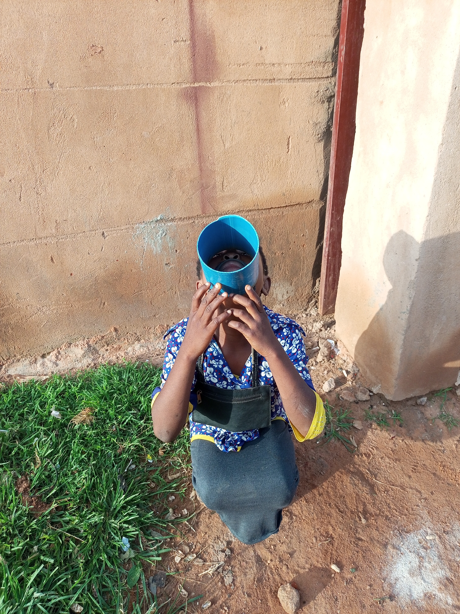 The image shows a person from the chest down, wearing a blue and white floral shirt with yellow trim and a dark skirt. They are holding a blue cylindrical object to their face, obscuring their eyes and nose. The person is seated on the ground in a rural or semi-rural area, with a patch of green grass to their left and a dirt ground to their right. Behind them is a textured, light brown wall. To the far right, a section of a lighter-colored wall is visible, with a vertical dark red post between the two walls. The scene appears to be outdoors during daylight. The location in Tempure, Nigeria, is not specified by any visible elements in the image.