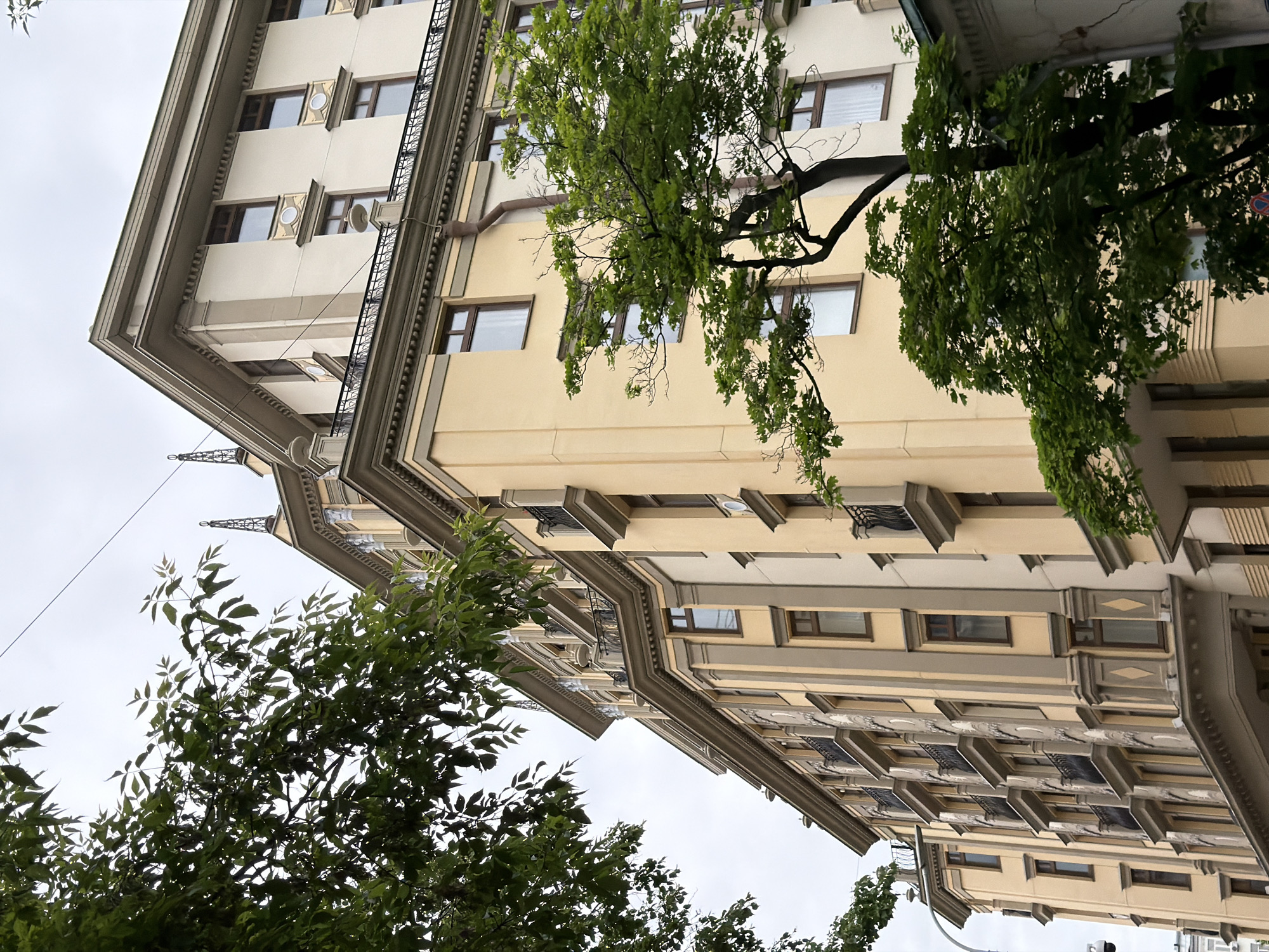 The image captures a low-angle, sideways view of two imposing, multi-story buildings, characteristic of urban architecture in Moscow. The primary building in the foreground, and a slightly receding one to its right, display facades primarily in a light cream or yellow hue, contrasted with prominent gray or beige architectural trim, cornices, and decorative elements. The style features strong horizontal lines, ornate window surrounds, and some windows are adorned with small, intricate wrought-iron balconies or grilles. Details such as circular decorative panels are visible above some windows on the left building.
Lush green foliage from several deciduous trees heavily frames and partially obscures the buildings, particularly on the lower left and upper right of the composition, indicating a spring or summer setting. A large tree branch extends across the upper mid-section of the image. The sky is overcast and light gray, suggesting a cloudy daytime.
No people or specific activities are visible, presenting a static architectural and natural landscape. A thin utility wire can be seen against the sky. A notable detail, partially hidden by foliage on the right building, is a red circular sign with a white diagonal bar, commonly indicating "no stopping." The sideways orientation of the photograph provides a dynamic and somewhat unconventional perspective of the urban environment.