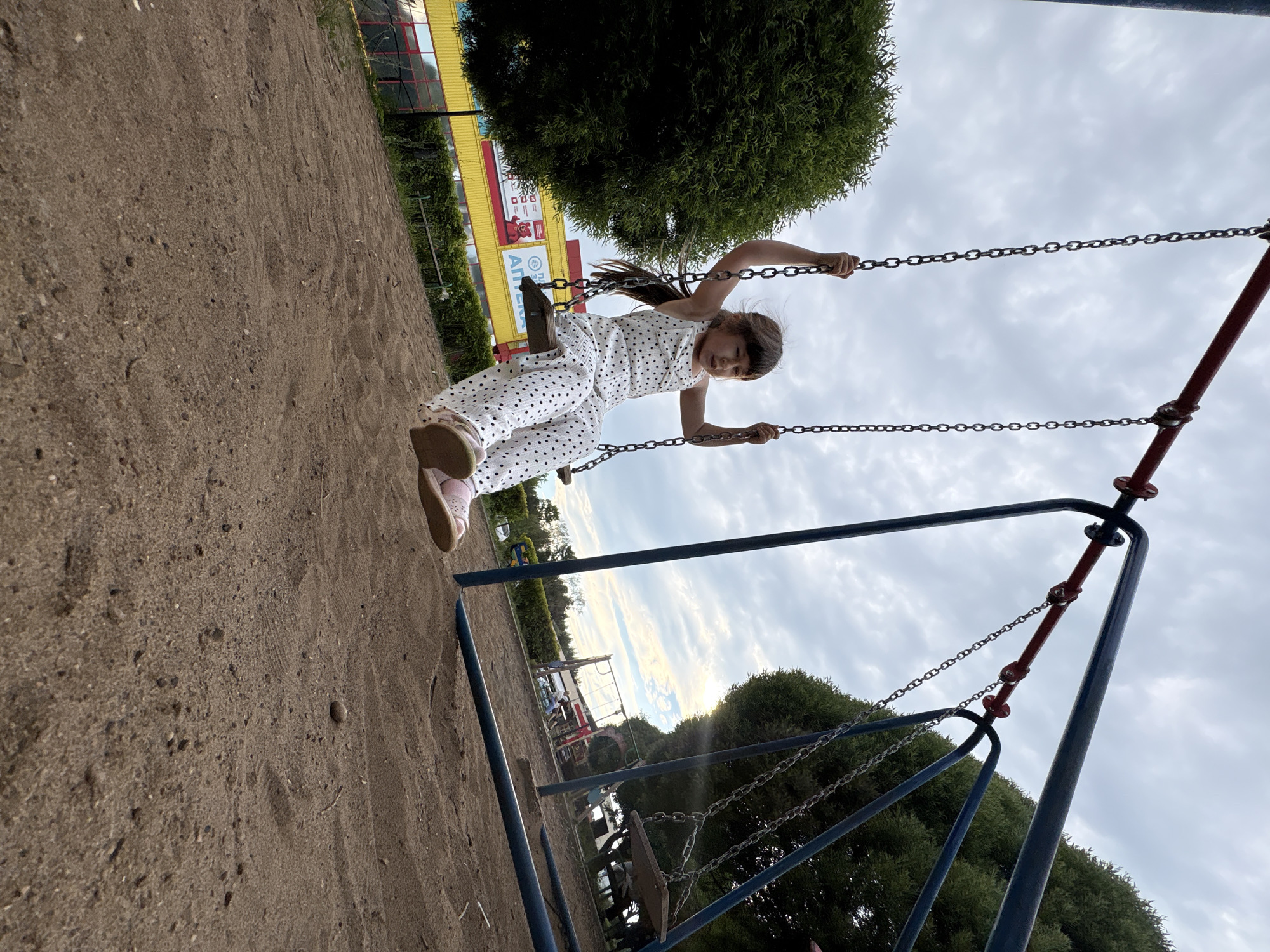 A young girl with dark hair is swinging on a swing set in a playground. She is wearing a white jumpsuit with black polka dots and pink sandals. The playground is made of sand and has green trees and a building in the background. The sky is overcast. The building in the background appears to be a store, with a yellow facade and red lettering. The lettering is in Russian and appears to be advertising an "APTEKA" which translates to "pharmacy." The playground is located in the city of Chaikovsky, Russia.
