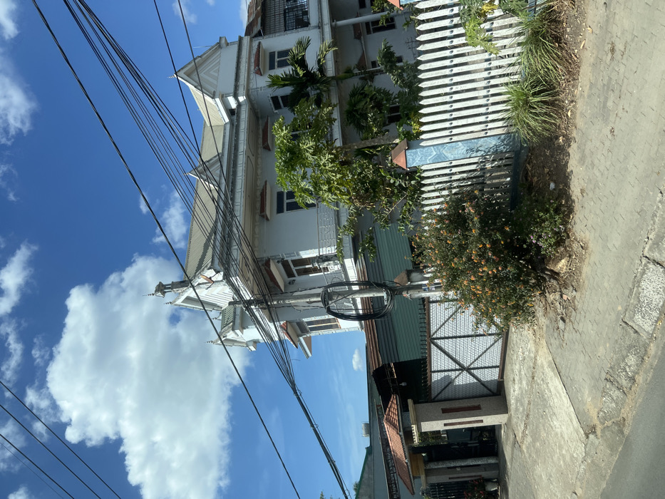 A white, multi-story building is viewed from a low angle under a bright blue sky with scattered clouds. Utility lines crisscross the sky. A white picket fence runs along the side of the building, bordered by green foliage and a paved sidewalk. Part of a gate and a smaller building with a red roof are also visible. The perspective is slightly tilted.