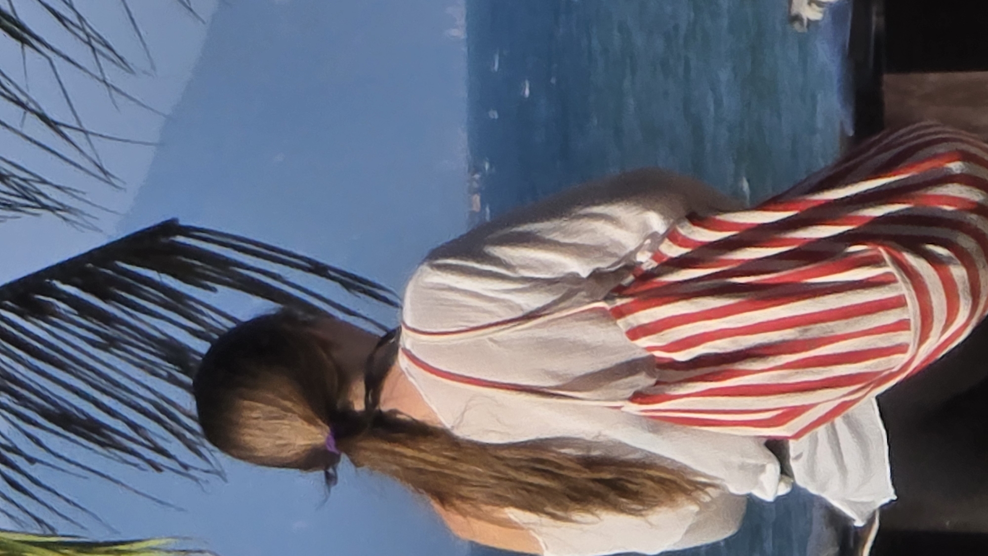 A person is seen from the back. They have long brown hair pulled back into a ponytail with a purple hair tie. The person is wearing a white shirt. The shirt has a thin red strap visible on the back of the neck. Over the shirt is a red and white striped bag or wrap.
In the background, there is a blue body of water, possibly the ocean. Behind the water is a hazy skyline, likely a city. There are also palm leaves visible in the left of the frame. The photograph was taken in Nha Trang Ward, Vietnam.
