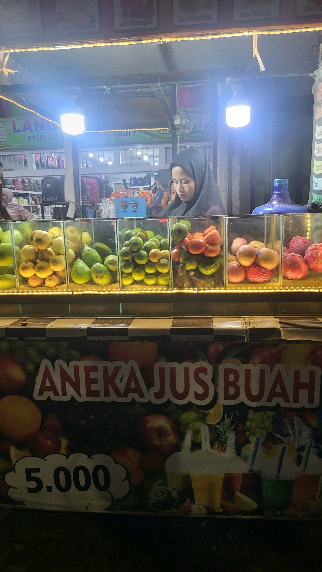 This image shows a nighttime scene at a fruit juice stand in Lebaksiu, Indonesia. A young woman wearing a black hijab is visible behind the counter, looking down at the fruit display. The stand features an assortment of fresh fruits in clear containers, including what appear to be oranges, avocados, limes, apples, and dragon fruit. Above the fruit display, signs indicate different juice flavors, such as "Jus Semangka" (watermelon juice) and "Jus Belimbing" (starfruit juice). A large banner below the fruit display reads "ANEKA JUS BUAH" (Various Fruit Juices) and shows a price of "5.000". Various bags and merchandise are visible in the background, suggesting the stand might be part of a larger market or shop. The lighting is artificial, with bright lamps illuminating the area.