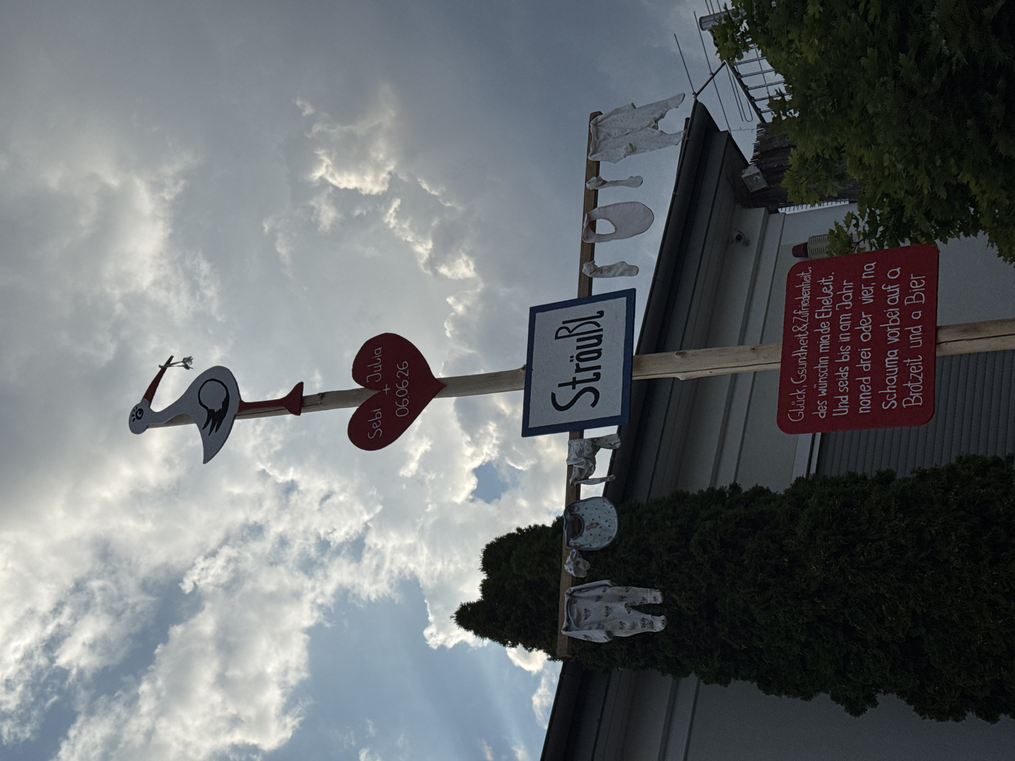 A wooden pole adorned with baby clothes and a stork decoration stands outside a building in Wettstetten, Germany. A red heart sign at the top of the pole reads "Seby + Julia 06.06.26" and a blue sign below it says "Sträußl". A red sign at the bottom of the pole contains a German poem. Baby clothes hang from a horizontal bar on the pole. The scene appears to be a celebration for a new baby. The sky is cloudy with rays of sunlight breaking through.