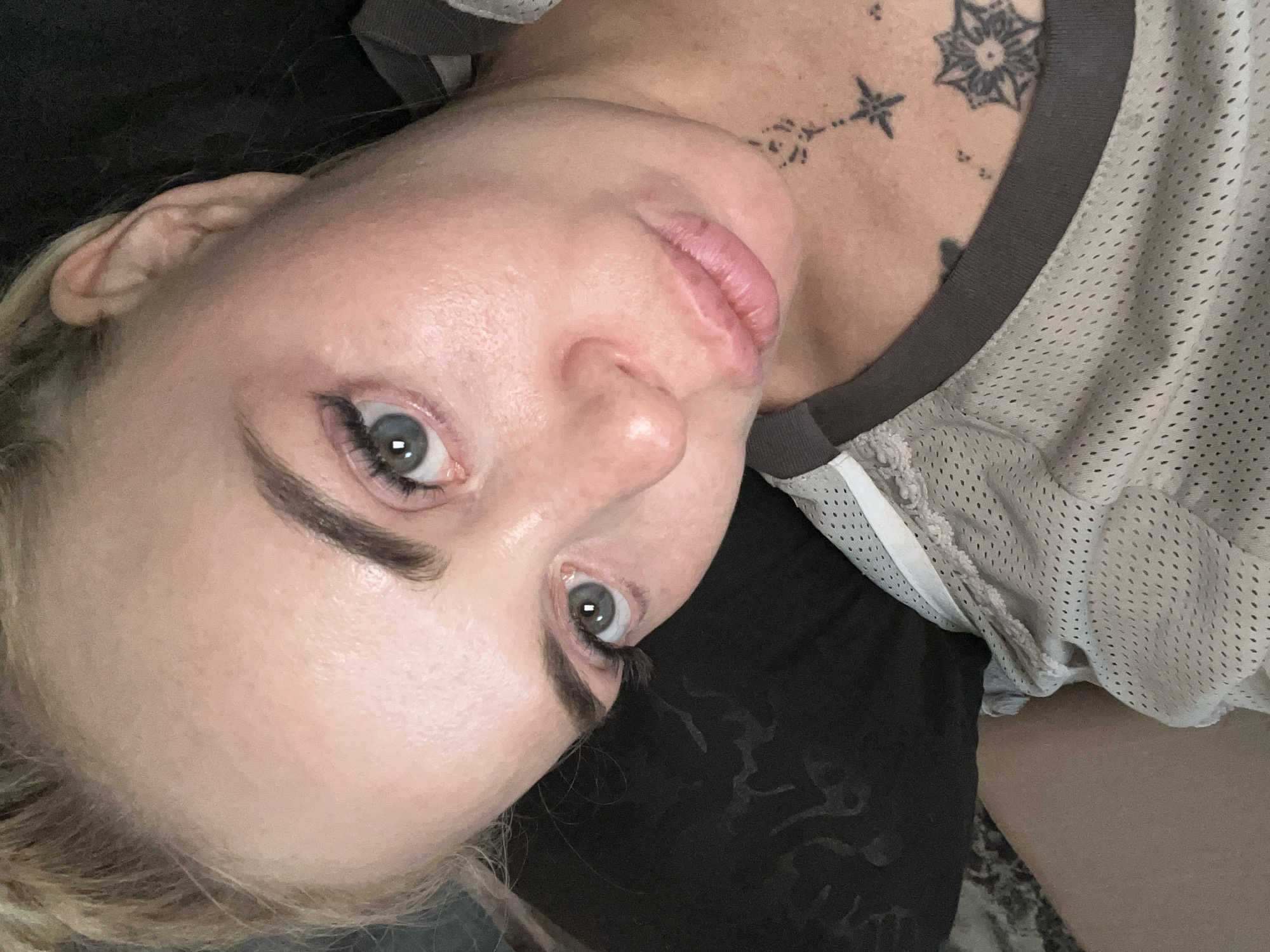 A close-up, overhead selfie features a woman with blonde hair pulled back, looking towards the camera with a neutral expression. Her face is illuminated by soft lighting, highlighting her pale skin, light grey eyes, and defined eyebrows. She is wearing a light-colored, mesh athletic top with grey trim. A tattoo of a star and a mandala design is visible on her upper chest. The background is dark and out of focus, with a hint of a patterned pillow or fabric on the left. The image is cropped to focus on the woman's face and upper torso.