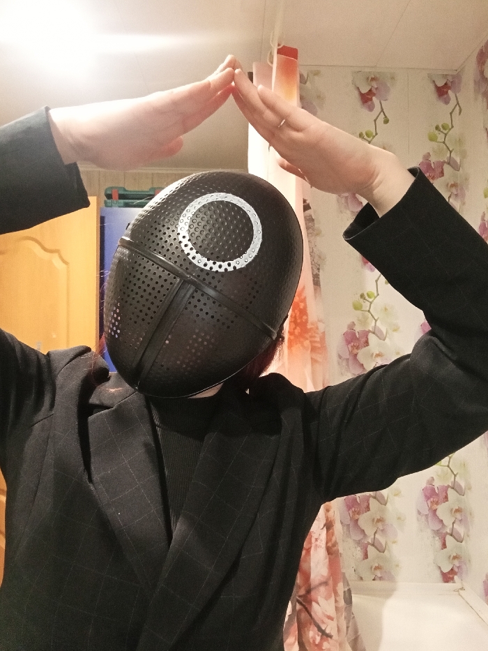 The image captures a person indoors, dressed in what appears to be a costume inspired by the Netflix series "Squid Game." The individual is wearing a distinctive black mask that completely covers their face. The mask has a textured, perforated surface with numerous small holes, and a prominent white circle symbol is painted on its forehead. The mask itself is segmented with dark lines, giving it a structured appearance.
The person is wearing a dark, possibly black or very dark gray, blazer or jacket over a dark turtleneck or high-collared garment. The blazer has a subtle, faint grid or checkered pattern visible across its fabric. Their hands are raised above their head, palms facing inward and slightly touching, forming an arched or roof-like shape over the top of the mask. A simple silver or white-gold band ring is visible on the ring finger of the person's right hand.
The background suggests an indoor setting, likely a room in a house or apartment. To the left, a light brown wooden door or cabinet is partially visible. The wall behind the person is covered with light-colored wallpaper featuring a floral pattern, specifically delicate orchids or similar flowers with light pink and white petals and green leaves, against a lighter background. Further back, on the right side of the frame, a vertical strip of orange or red material, possibly a curtain or part of another object, can be seen. The lighting appears to be artificial and warm, illuminating the scene from above. The overall impression is a posed shot, possibly for a costume display or a casual photo.