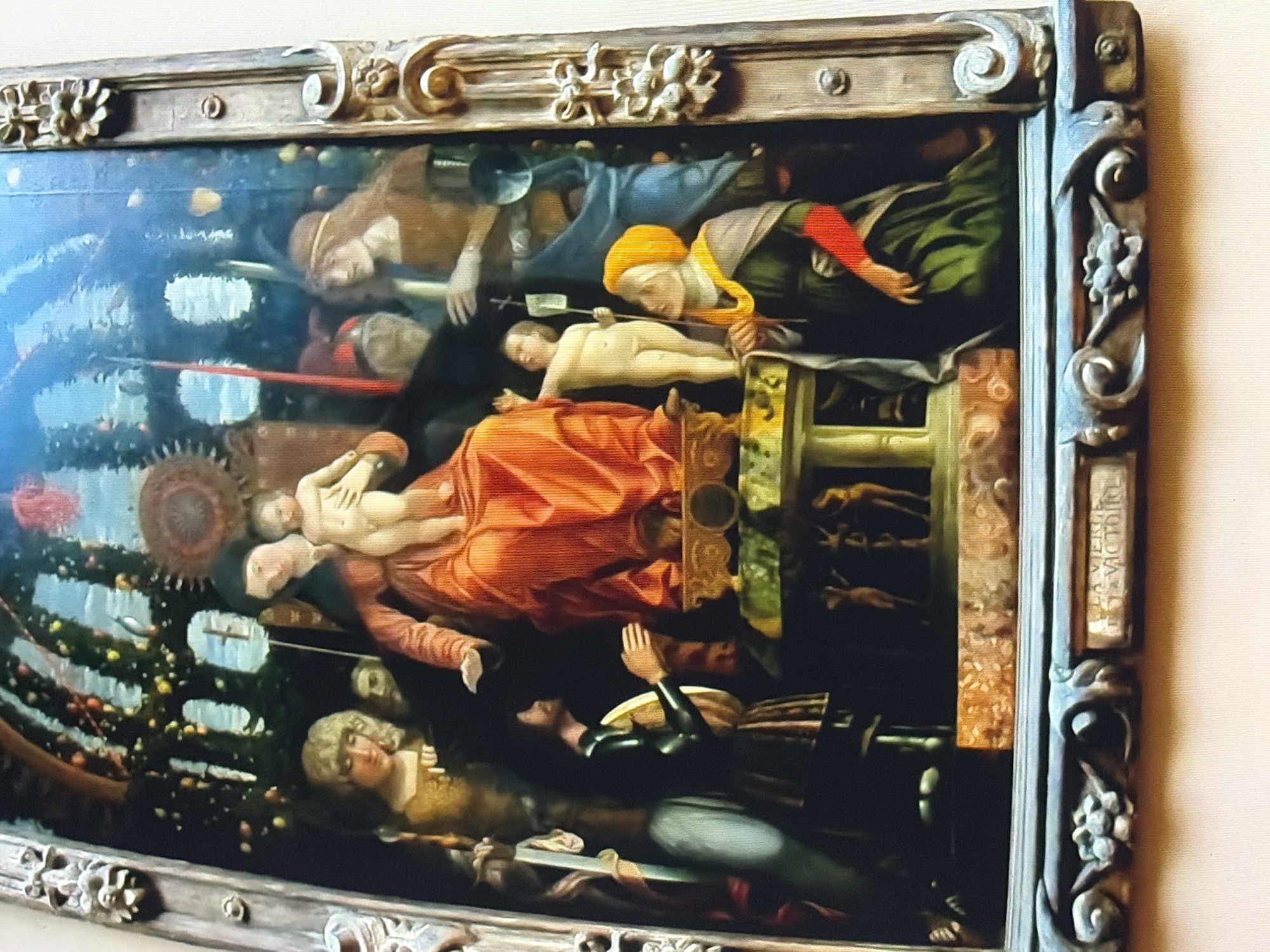 A richly detailed religious painting depicts the Madonna and Child enthroned, surrounded by a retinue of figures. The central figures are a woman in an orange-red robe and a cherubic infant held in her arms. She has dark hair and is seated on a raised platform adorned with intricate carvings and a green base. The child, naked, reaches out with one hand. To the left of the Madonna, a woman with blonde curly hair gazes downwards. Behind her, two male figures are visible, one with a dark cap. To the right of the central group, a woman in a green gown and a yellow headscarf looks upwards with her hands clasped. Her left sleeve is a striking red. A man in dark attire with his hands clasped in prayer stands to the left, facing away from the viewer. The background suggests an architectural setting, possibly an archway adorned with greenery and fruits, with a bright blue sky visible beyond. The entire scene is framed by an ornate, dark wooden border with sculpted floral motifs. At the bottom of the frame, a rectangular plaque bears the inscription "LA VIERGE ET LA VICTOIRE." The painting appears to be displayed in a museum setting, with a pale wall visible in the background.