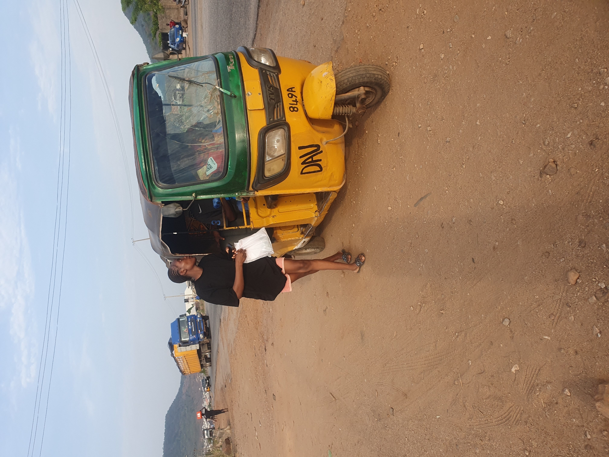 A woman stands on a dirt road in Lokoja, Nigeria, next to a yellow and green auto rickshaw, holding a white plastic bag. The rickshaw has the word "DAV" painted on its side and a license plate that reads "849A." The woman wears a black shirt, pink shorts, and sandals. In the background, a blue and yellow truck is parked along the road, and a hill rises in the distance. Overhead power lines are visible against a pale sky. The road surface is uneven with gravel.