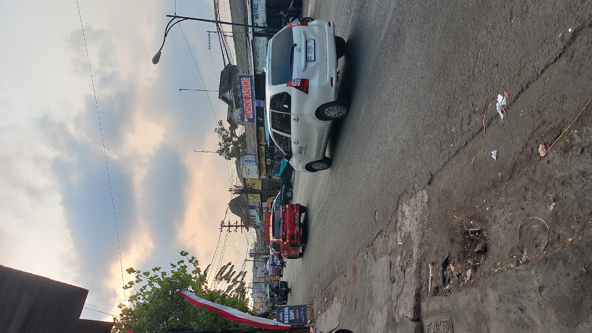 The image shows a street scene in Mangunsari, Indonesia, with several cars and motorcycles on the road. The sky is overcast with scattered clouds. On the left side of the road, there is a sidewalk with some debris. A sign on the left reads "JUAL DAGING SAPI" (Beef for Sale). There are buildings lining the right side of the road, some of which appear to be shops or restaurants. One sign on the right reads "MASAKAN PADANG" (Padang cuisine). A white van with the logo "ELGA" is visible in the foreground, and a red car is in the middle distance. The overall impression is a typical street scene in an Indonesian city with moderate traffic and commercial activity.