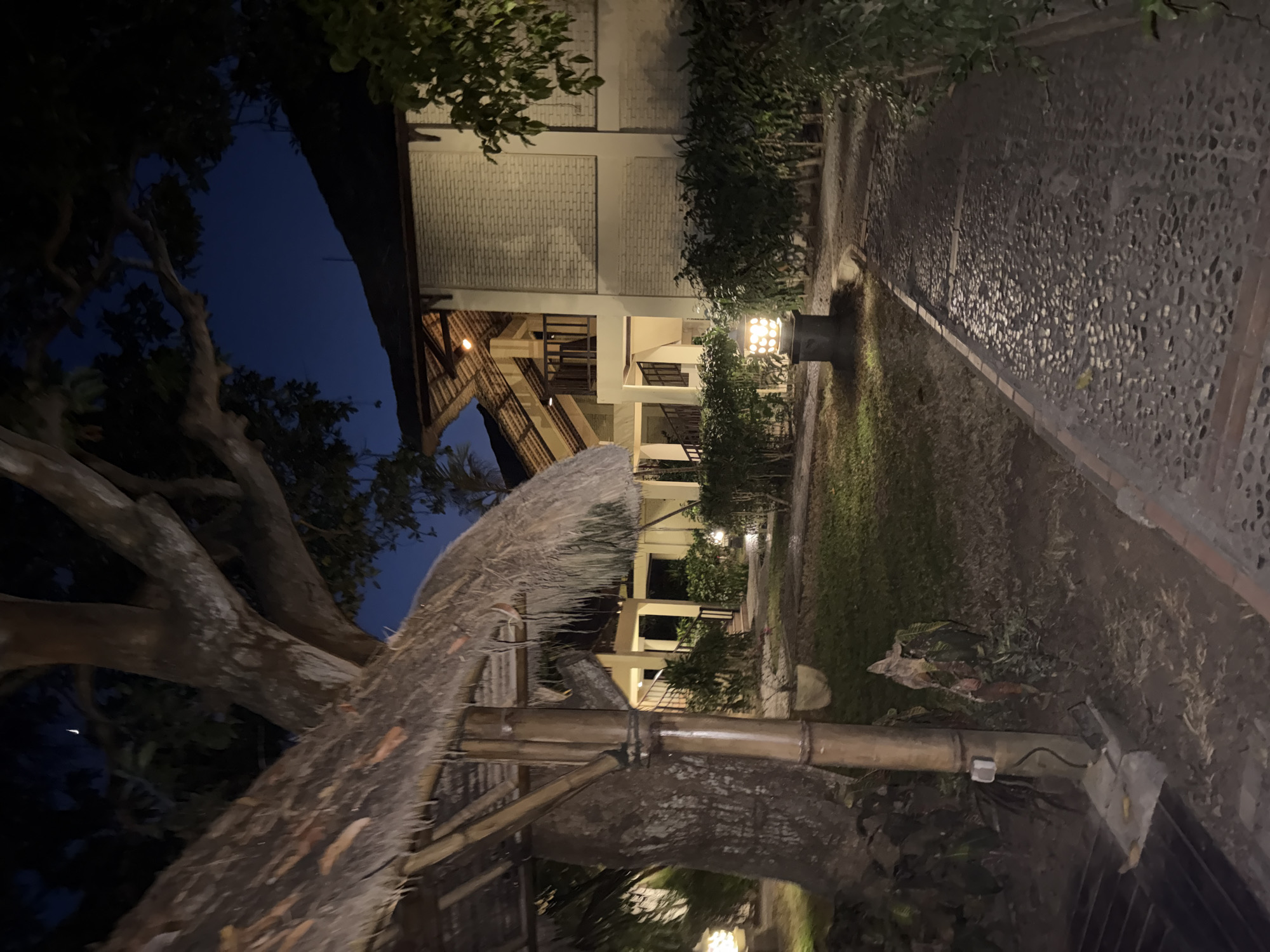 A nighttime exterior scene depicts a pathway paved with small stones leading through a garden area. A thatched-roof gazebo with bamboo supports is visible on the left, adjacent to a large tree. In the background, a multi-story building with a pitched roof and balconies is illuminated. Several outdoor lamps are positioned along the pathway and within the garden. The sky above is dark blue, with a faint crescent moon visible. The environment appears to be a resort or hotel in the city of Denpasar, Indonesia, given the architectural style and tropical foliage. There are no people or animals present in the image.