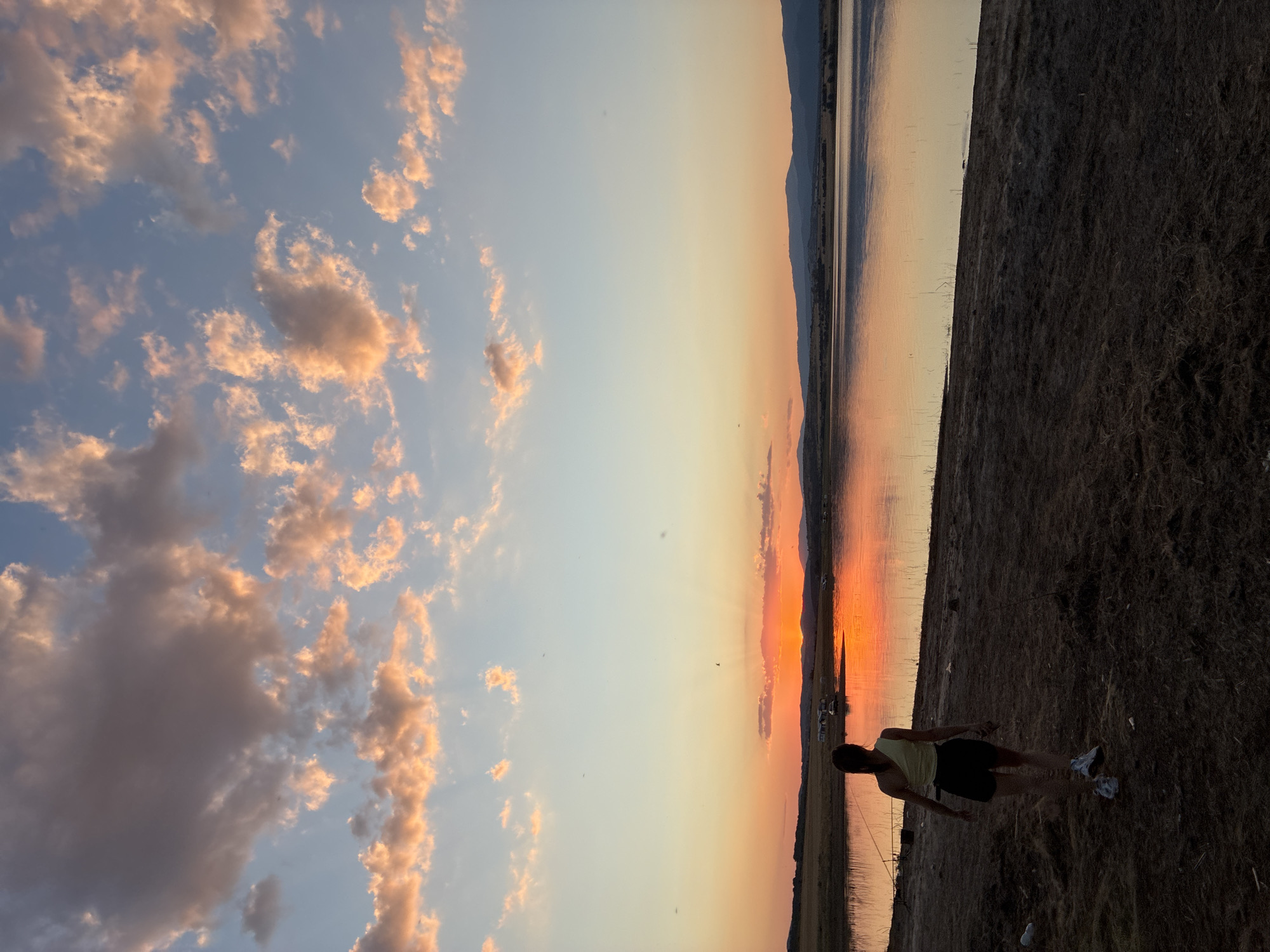 A young woman with dark hair, wearing a yellow top and black shorts, walks along a dirt path next to a body of water. The sun is setting, casting a warm orange and pink glow across the sky and reflecting on the water. In the distance, mountains are visible on the horizon. The environment appears to be a natural landscape, possibly a reservoir or lake, with dry vegetation on the shore. The lighting suggests it is either early morning or late evening.