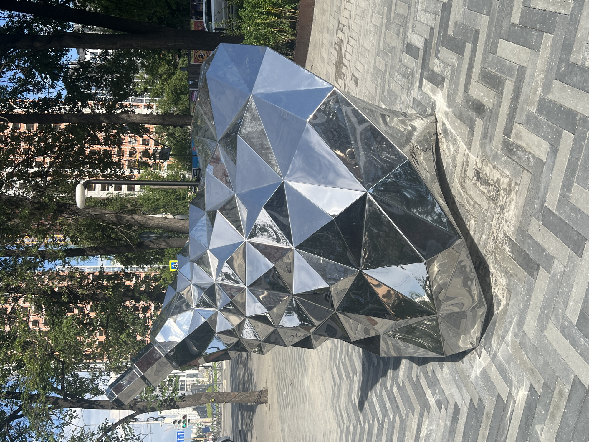The image shows a futuristic, geometric sculpture made of polished metal, placed on a patterned sidewalk in Yekaterinburg, Russia. The sculpture is a large, abstract form with many facets, reflecting the surrounding environment of trees, buildings, and a street with traffic signals. The sidewalk is paved with grey and black rectangular bricks laid in a herringbone pattern. In the background, there are tall trees with green leaves, modern buildings with orange and white facades, and a clear blue sky. A street lamp is visible to the left of the sculpture. There are no people visible in the immediate vicinity of the sculpture. The lighting suggests it is a bright, sunny day.