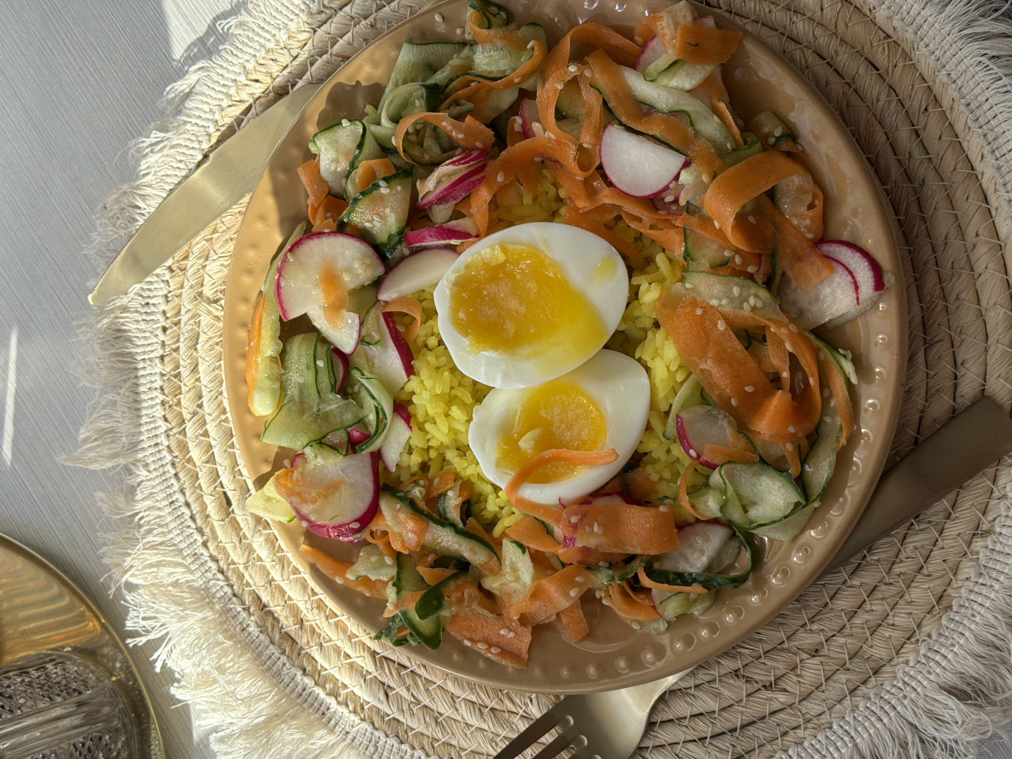 A top-down, well-lit shot showcases a plate of food on a natural-fiber placemat. The plate is filled with yellow rice topped with a halved soft-boiled egg. Around the rice and egg, there's a mix of thinly sliced vegetables, including carrots, cucumbers, and radishes, seemingly dressed with sesame seeds. The scene includes a gold-colored knife and fork flanking the plate. The food and tableware rest on a white surface, possibly a table. The location of Krasnoyarsk, Russia, is not ascertainable from the image.