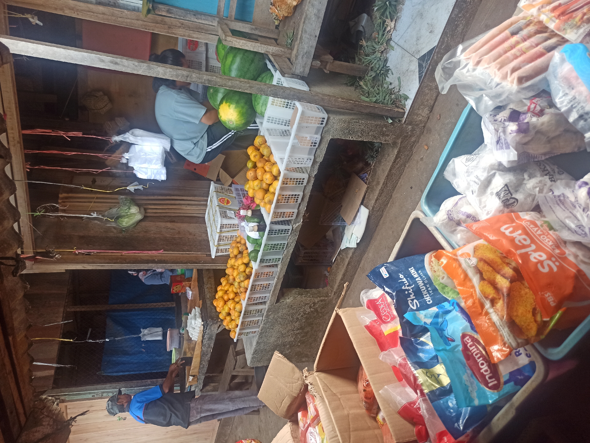 This image depicts an outdoor market stall in Trisinar, Indonesia. The stall appears to be selling fruits, vegetables, and packaged food items.
In the foreground, several large cardboard boxes are stacked, filled with various packaged food products, including what appear to be frozen snacks and processed meats. One box in the lower left contains an assortment of colorful, sealed bags, some with prominent branding like "Indomina" and "Shifudo." Another teal plastic bin in the foreground is filled with more packaged food, including a bag labeled "Salam" with an image of fried food items and "Bakso Ayam." To the right of this bin, another plastic container holds numerous sausages in clear packaging, alongside bags of other frozen goods.
In the middle ground, a table or counter displays a large quantity of oranges piled high in white plastic crates. Beside the oranges, there are more white crates, some containing green produce, possibly avocados or limes, and a bottle of liquid. Further back, a person in a light blue t-shirt and dark pants is seen from the side, appearing to be attending to customers or organizing goods. Behind them, a second person in a light gray t-shirt is bending down, possibly reaching for items. The background reveals the open structure of the market stall, with wooden beams and a thatched or corrugated roof. Blue tarps are visible behind the stall, suggesting it is part of a larger market area.
The environment is rustic, with the wooden construction of the stall and the presence of loose vegetation and debris on the ground. The overall impression is a busy, functional market setting.
