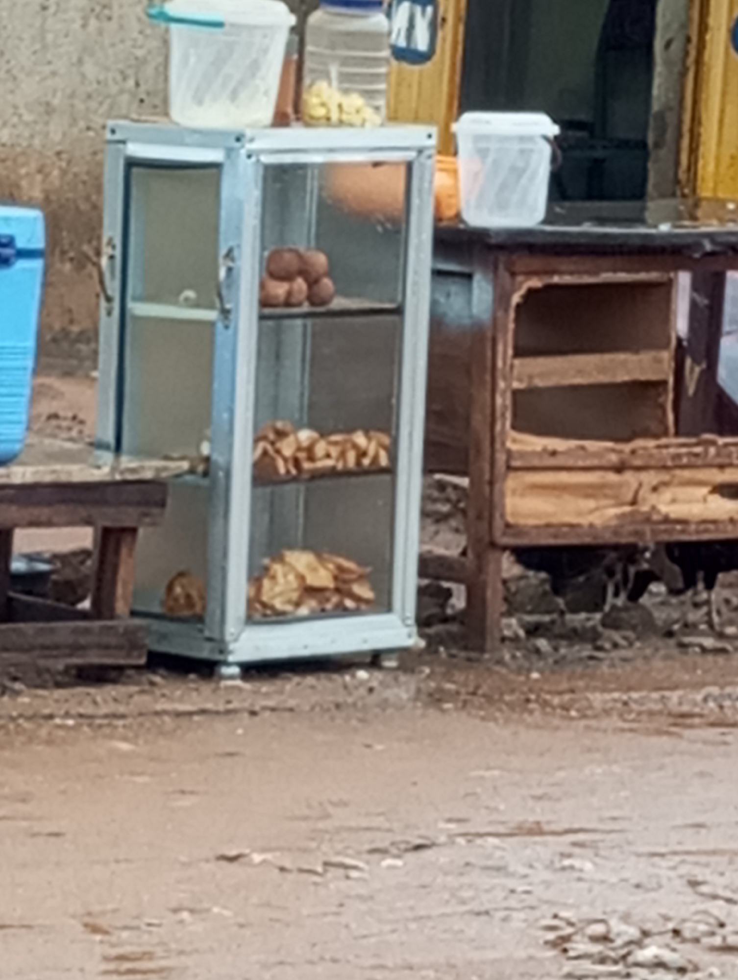 The image shows a street scene in Sabon Tasha, Nigeria. A glass display case containing bread or pastries sits on a small table. A second, larger wooden structure with shelves is next to it. Two white plastic containers are on top of the display and wooden structure. The ground is cracked and uneven. Several chickens are visible underneath the wooden structure.
