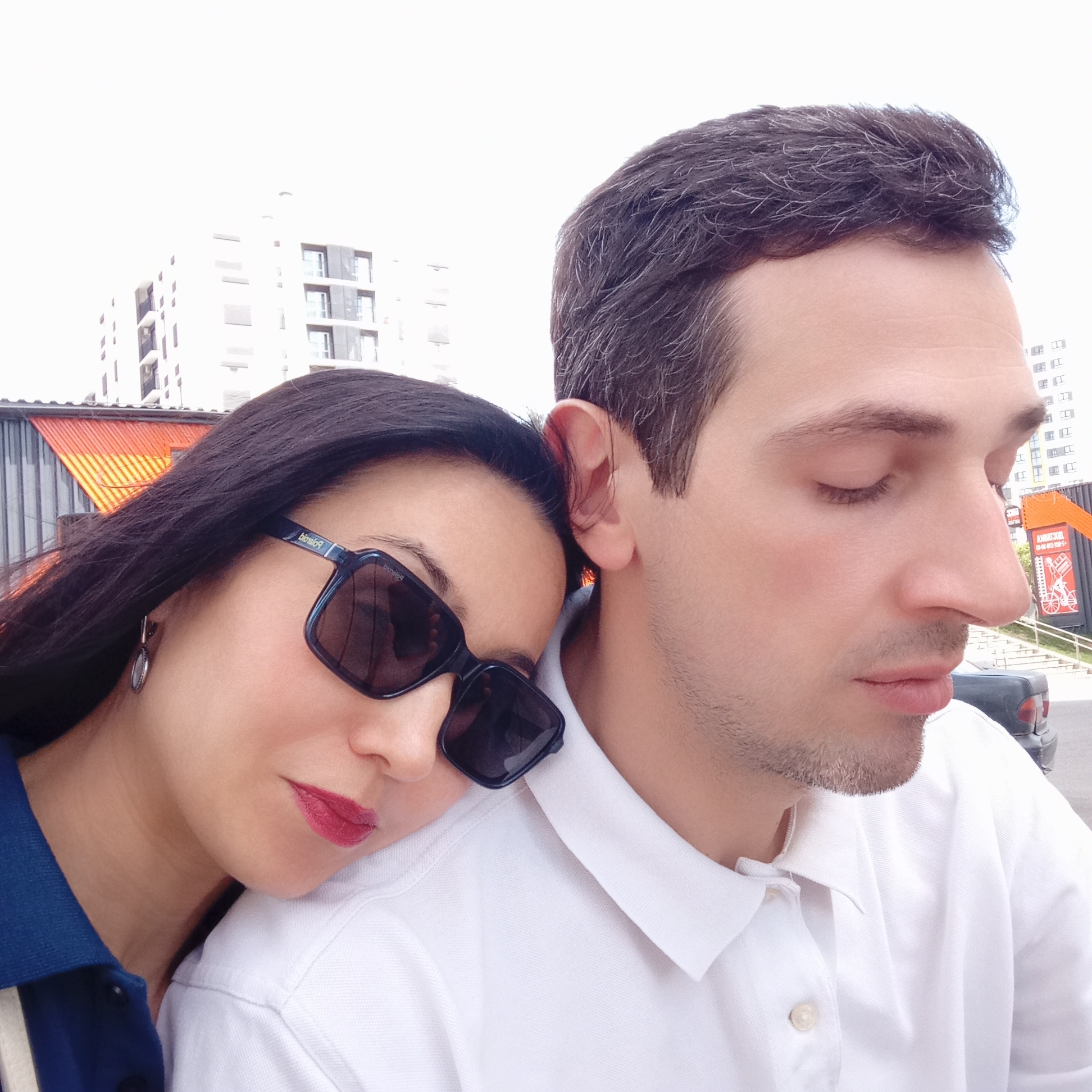 A medium shot shows a woman and a man close together in an outdoor setting. The woman is on the left, wearing dark sunglasses and dark lipstick. Her head rests on the man's shoulder. The man is on the right, eyes closed. They both wear polo shirts; the woman’s polo shirt is blue with white stripes. The man’s polo shirt is white.
In the background are buildings and other urban elements. Orange roofs of what appear to be commercial buildings are visible. Behind them, a taller, white apartment building with multiple windows rises. A dark-colored car is partially visible behind the man. A promotional poster is also visible in the background with red text.
The location is Pervomaysky, Russia.