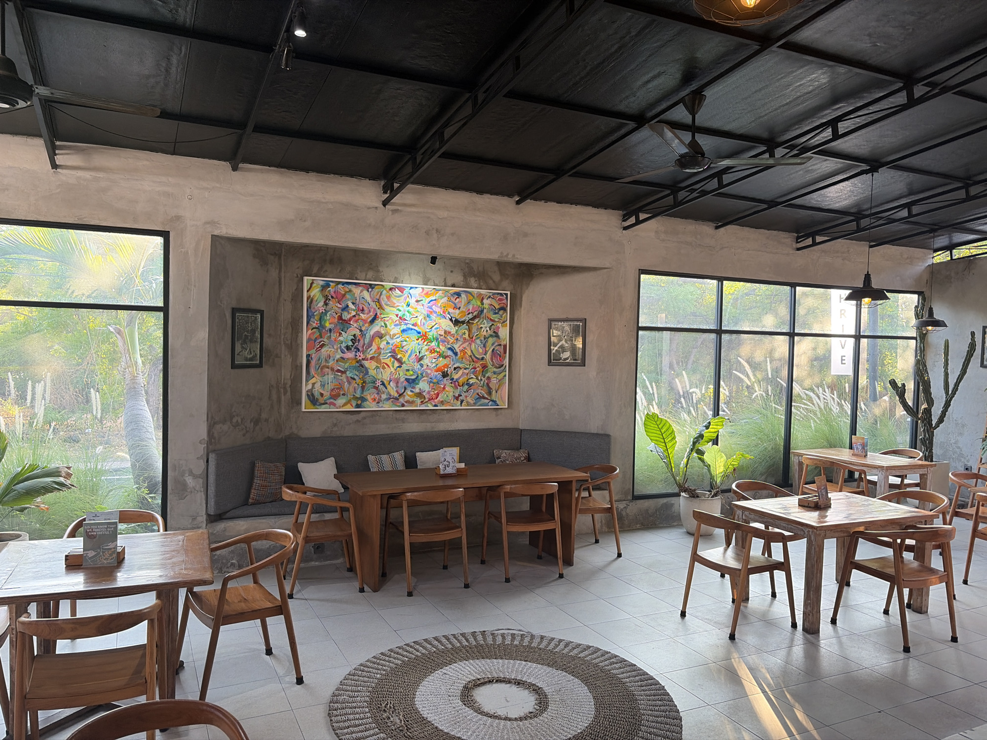 The image shows the interior of a cafe or restaurant with an outdoor seating area visible through large windows. The cafe has a modern industrial design with exposed metal beams on the ceiling and concrete walls. Several wooden tables and chairs are arranged throughout the space, with some set up for dining and others as lounge seating. A large, colorful abstract painting hangs on the wall above a built-in bench, which is adorned with decorative pillows. Potted plants are placed strategically, adding a touch of greenery to the minimalist decor. The lighting appears to be a mix of overhead industrial pendant lamps and natural light streaming in from the windows. In the background, through the windows, the scene transitions to an outdoor environment with lush vegetation and tall grasses, suggesting a tropical or semi-tropical location. The floor is tiled with a light-colored material, and a circular woven rug is placed in the foreground. The overall impression is one of a relaxed and stylish dining or gathering space.
Based on the context provided by the user, this scene is located in the city of Kubu, Indonesia.