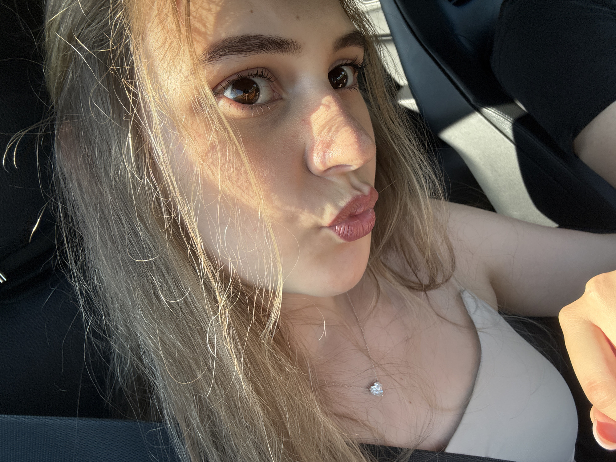 This media file is a close-up selfie of a young woman with long, blonde hair. She is wearing a light-colored tank top and a delicate silver necklace with a heart-shaped pendant. Her facial expression is a playful pout, with her lips pursed. The lighting suggests it is daytime, with sunlight casting shadows across her face and hair, creating a slightly dramatic effect. The background appears to be the interior of a car, with dark upholstery visible, indicating she is likely a passenger or driver. The image captures a casual and spontaneous moment, possibly taken while out and about in Iffezheim, Germany. There is no visible text in the image.