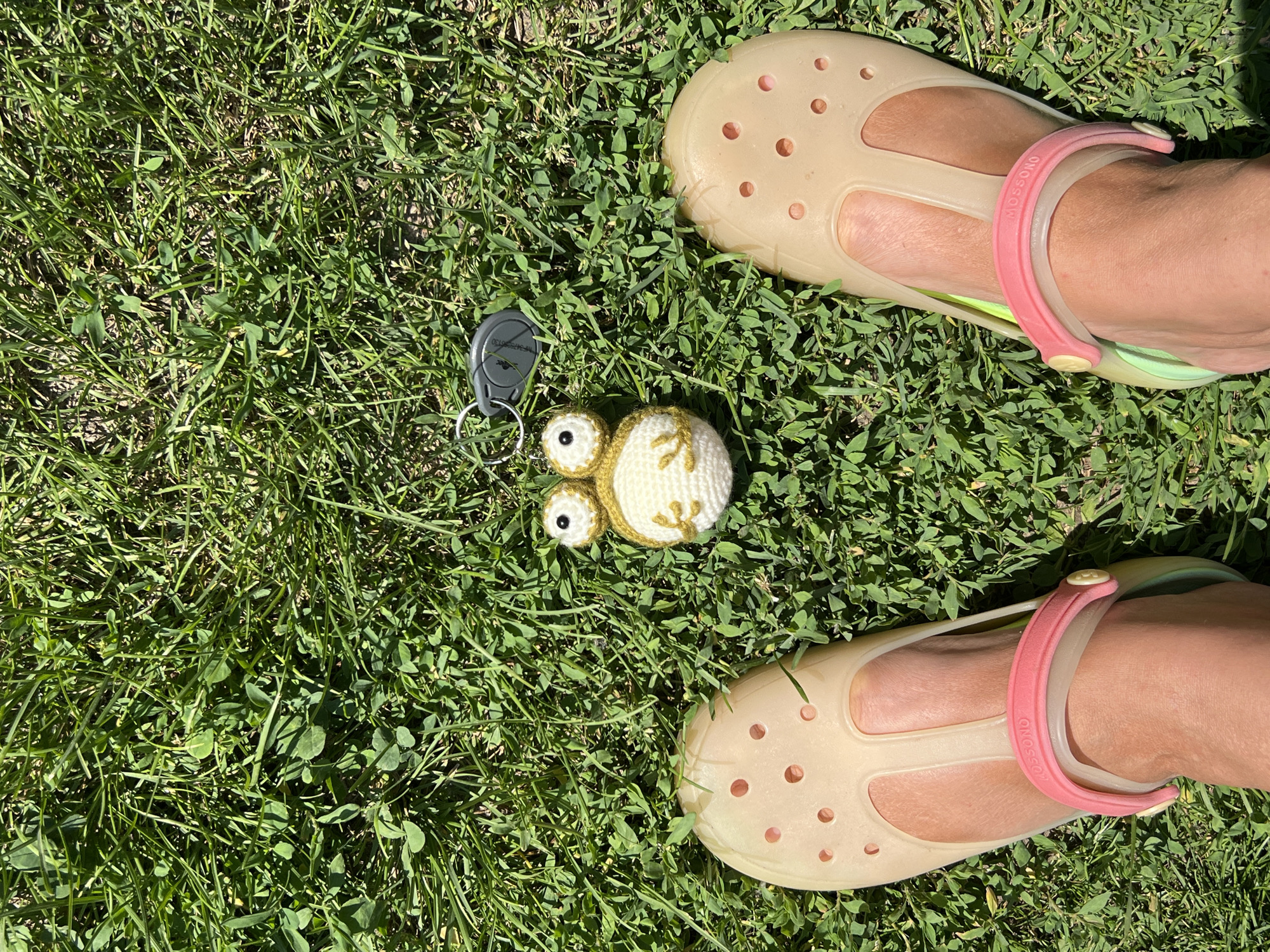 A top-down, full-frame shot captures a patch of green grass bathed in sunlight. Two feet, adorned with light tan crocs and pink straps, are positioned towards the bottom of the frame. Between the feet, a small crocheted frog keychain with big eyes and a white body rests on the grass, next to a grey car key fob. The scene is set outdoors on a sunny day. The location is identified as Mochische, Russia.