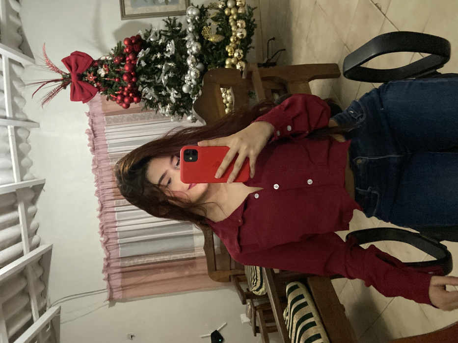 A woman sits sideways in a black chair, taking a selfie with a red phone. She wears a long-sleeved maroon shirt and blue jeans. Behind her, a decorated Christmas tree with red and silver ornaments sits near a window covered by a pink curtain. The reflection in a mirror shows part of the room including a white-slatted structure. The image is taken indoors with soft lighting.