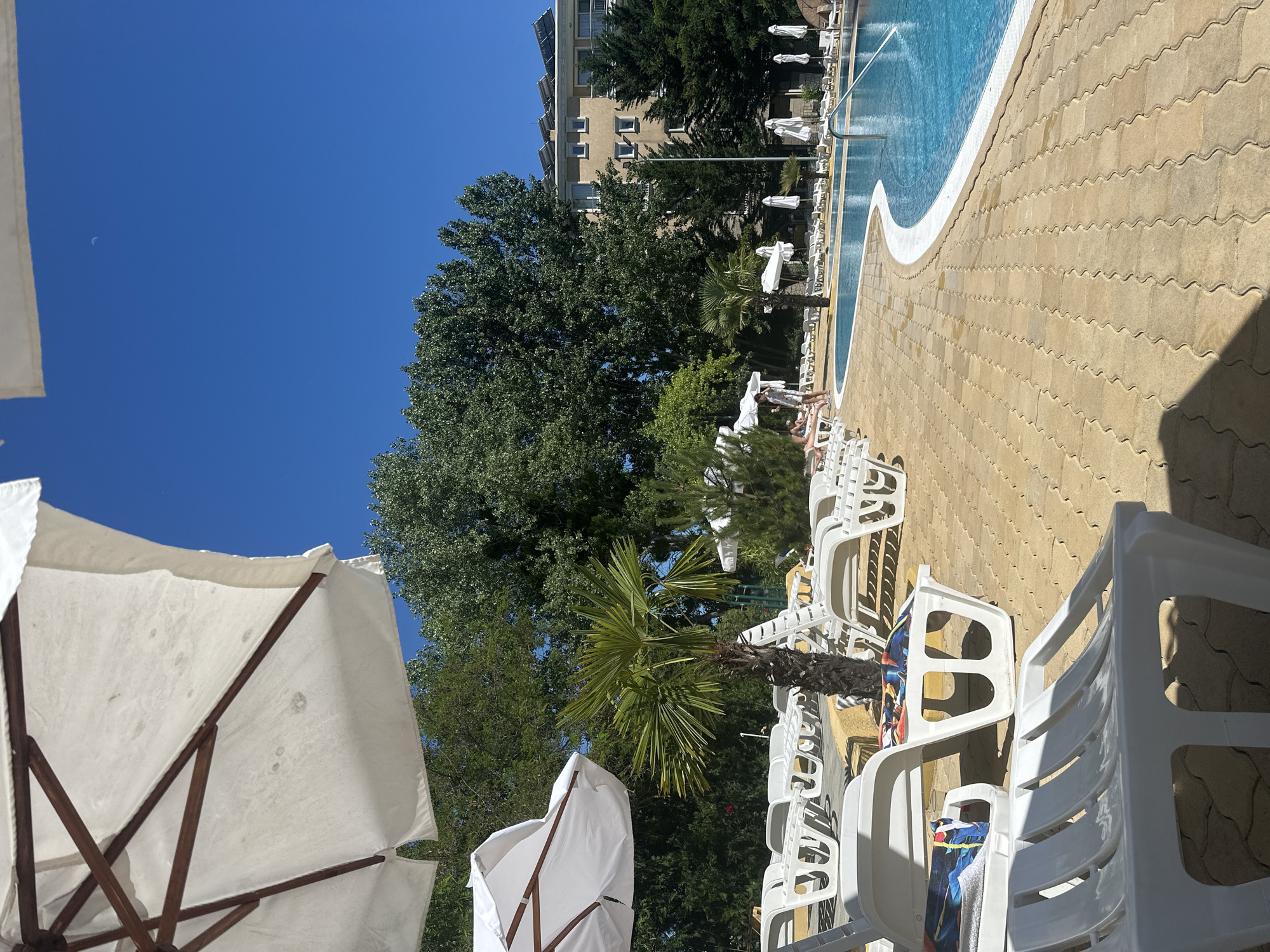 The image shows a sunny day at a resort pool, likely in Nesebar, Bulgaria. In the foreground, several white plastic lounge chairs are arranged on a yellow paved area, some with colorful towels on them. A large white umbrella provides shade in the upper left corner. A small palm tree stands behind a row of lounge chairs, and beyond that, more lounge chairs are visible, with a few people relaxing on them. A swimming pool with bright blue water is to the right, bordered by a white edge. In the background, a multi-story building with solar panels on the roof is surrounded by lush green trees. The sky is a clear, bright blue.