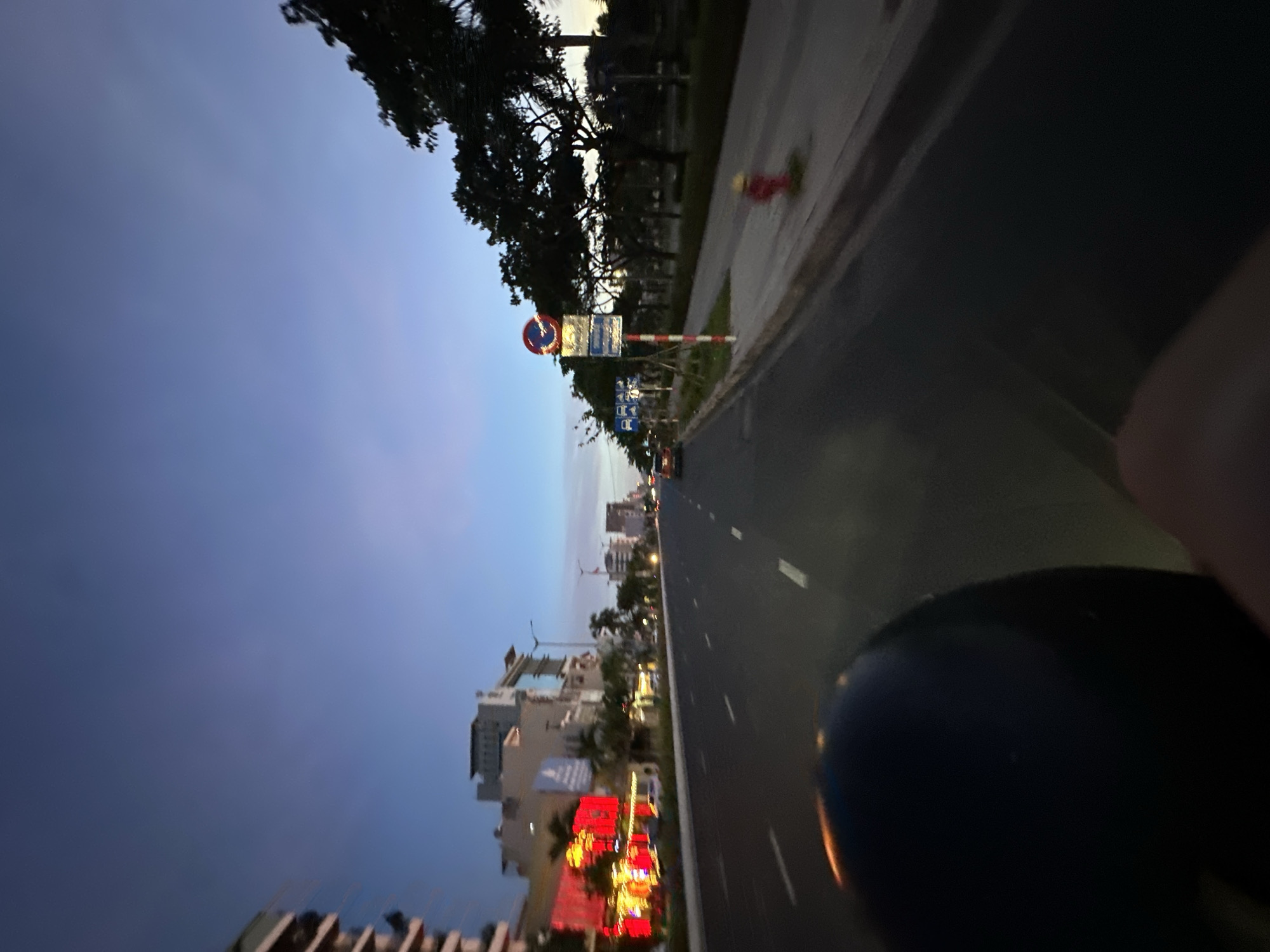 The image captures a perspective from a moving vehicle, likely a motorcycle, on a wide, paved road in Da Nang, Vietnam. The time of day appears to be twilight or early evening, as indicated by the deep blue and violet hues of the sky. The road features white dashed lines marking lanes.
On the left side of the road, there are multi-story buildings with illuminated signs, some displaying bright red and yellow lights, suggesting commercial establishments. Palm trees are visible interspersed between the buildings.
On the right side of the road, a sidewalk runs alongside a row of trees. A red fire hydrant is positioned near the sidewalk's edge. Several traffic signs are mounted on poles along the sidewalk. One prominent sign, circular with a red border and a diagonal red line across a blue background, indicates a prohibition. Another rectangular sign below it provides further information, likely related to traffic regulations. A few other signs with blue backgrounds and white symbols are also visible, some possibly indicating parking restrictions or information for drivers. The overall impression is of a well-developed urban environment.