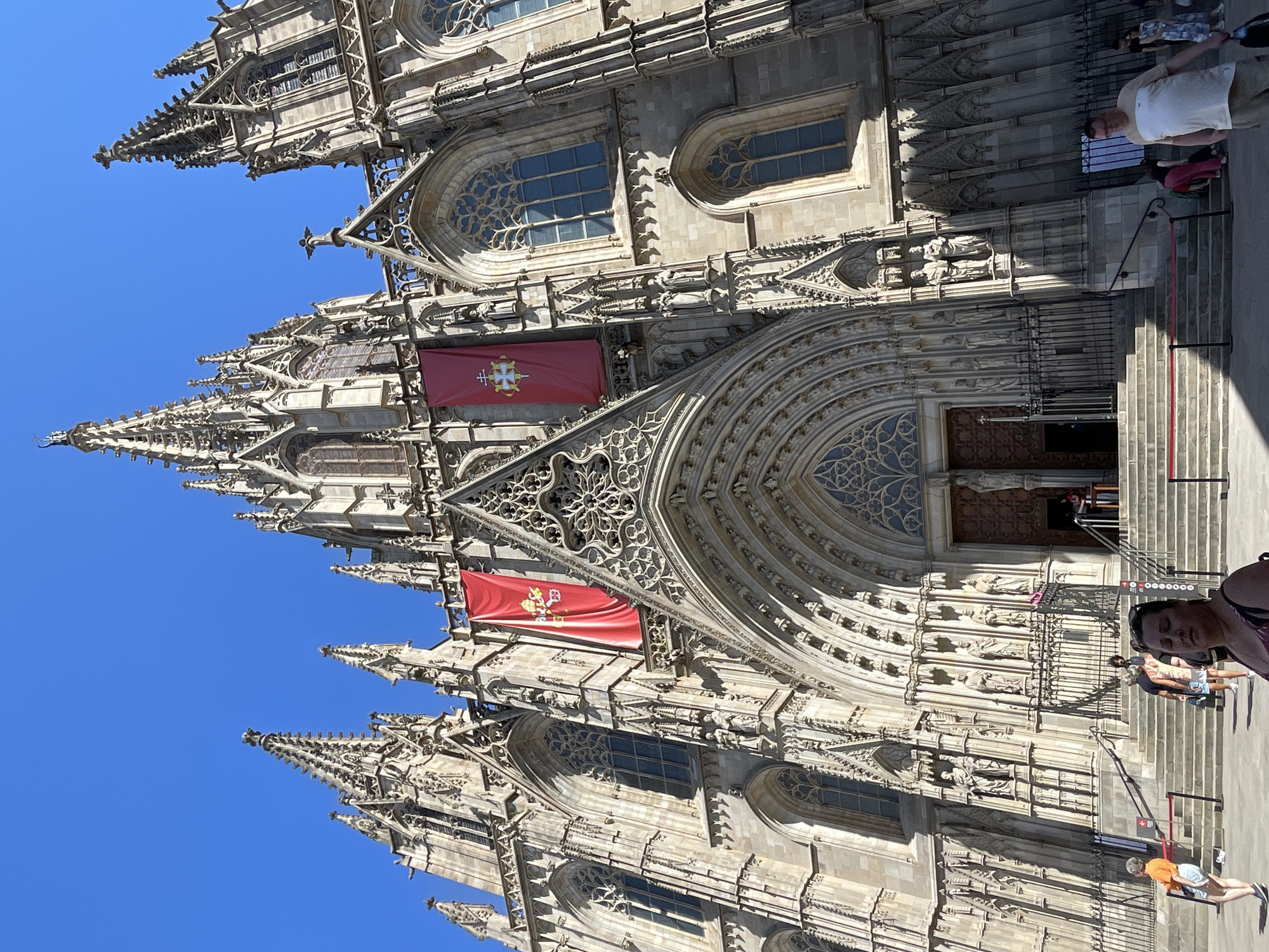 The image shows a low-angle view of the Barcelona Cathedral (Catedral de Barcelona) in Barcelona, Spain, under a clear blue sky. The cathedral's facade is made of stone and features intricate Gothic architecture, including pointed arches, spires, and ornate carvings. Two large red banners with gold emblems are hung on either side of the main entrance.
In the foreground, several people are visible. On the left, a woman in an orange shirt and white shorts walks towards the cathedral steps. In the center foreground, a woman with dark hair and a patterned top is looking downwards. To the right, a man in a light-colored t-shirt and khaki shorts walks away from the camera. Other individuals are seen walking up the stairs or standing near the entrance.
The entrance to the cathedral has a large, ornate wooden door, and a set of wide stone stairs leads up to it. Red velvet ropes are strung along the stairs, seemingly directing foot traffic or marking an entrance. The overall scene depicts a sunny day with tourists and visitors at a prominent religious landmark.