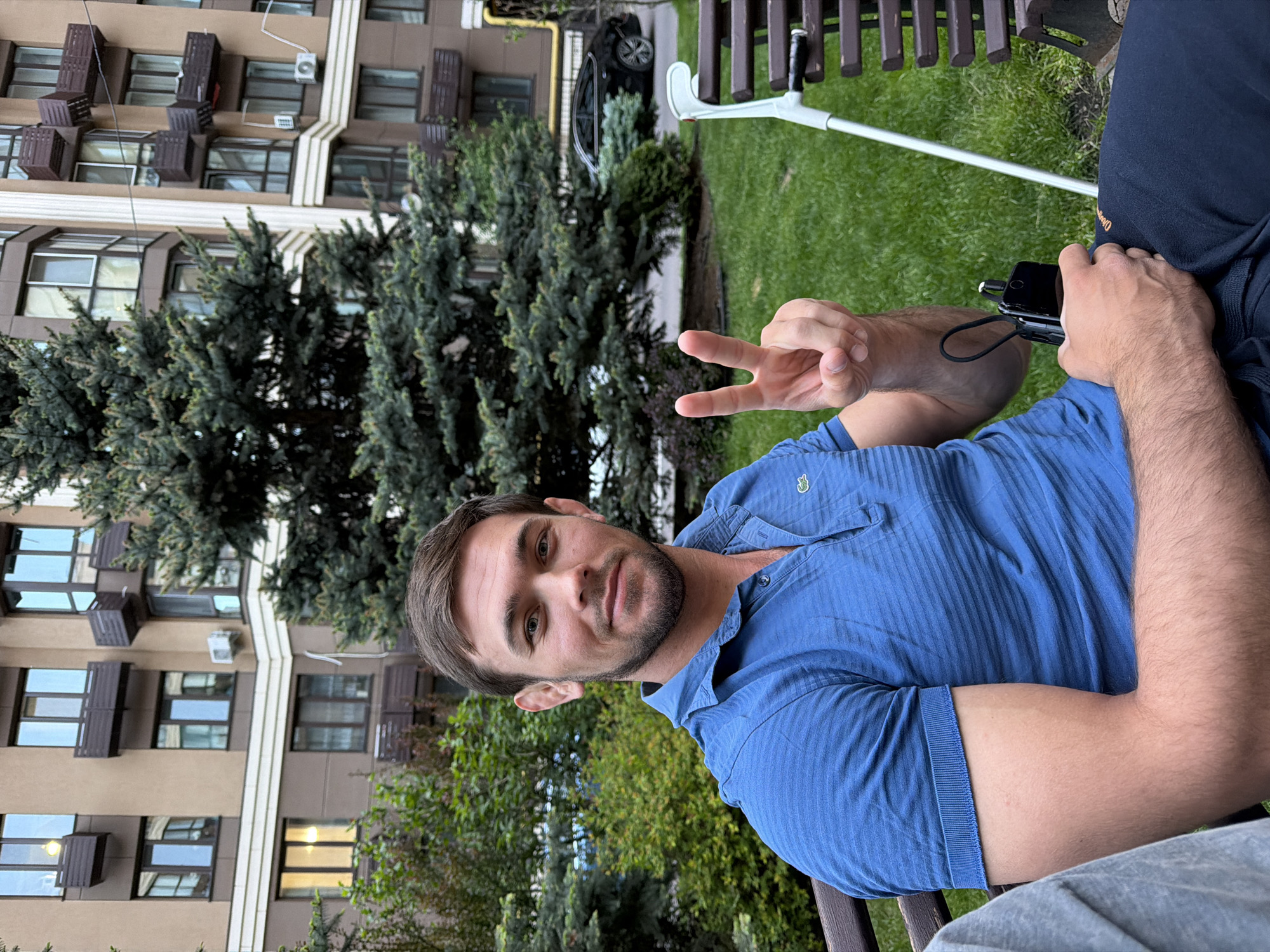 A man is captured in a horizontal portrait, seated outdoors in a residential setting in Sofiivska Borshchahivka, Ukraine. He has short brown hair and a beard, and wears a blue polo shirt with a small, possibly crocodile, logo on the left chest, and dark blue shorts or trousers with some faint, illegible white text. He looks directly at the camera with a calm, relaxed expression, making a peace sign with his left hand. His right hand holds a black smartphone, which has a black lanyard attached.
Beside the man, a white and black crutch rests on the vibrant green grass. The background features several large, dark green coniferous trees, and behind them, a modern, multi-story residential building with brown and cream-colored facades and numerous windows. Some windows reflect the daytime light, and several external units or decorative elements are visible on the building. A black car is partially visible parked on a paved area further back. The scene suggests a pleasant, possibly late afternoon, atmosphere.