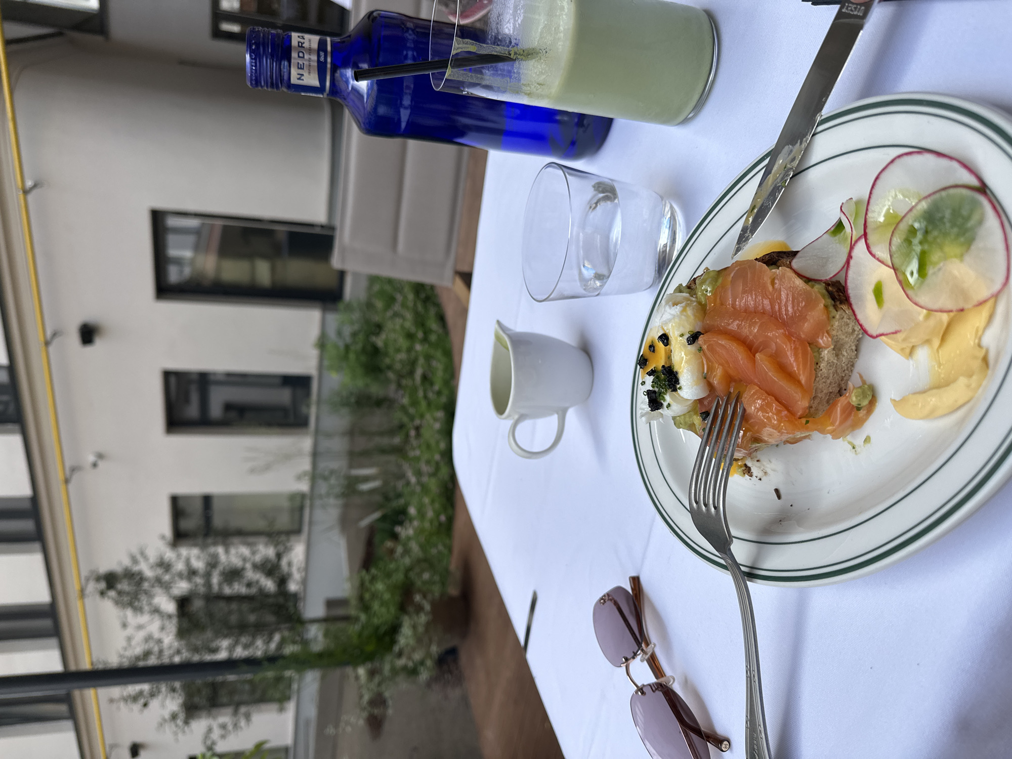 A still life scene on a white tablecloth depicts a breakfast spread at an outdoor cafe in Saint Petersburg, Russia. The foreground features a white plate with a partially eaten open-faced sandwich topped with smoked salmon, avocado, a poached egg, and black caviar. Thinly sliced radishes and a dollop of yellow sauce are also on the plate, with a fork and knife positioned for eating. To the left, a pair of rose-tinted sunglasses rests on the tablecloth. In the background, a blue bottle of "NEDRA" water stands next to a glass containing a green beverage with a straw. A small white creamer jug is also present. The cafe appears to be situated in an courtyard, with a building in the background featuring white walls and dark windows. Green foliage and plants are visible in the mid-ground, suggesting a pleasant outdoor dining atmosphere. The lighting indicates it is daytime.