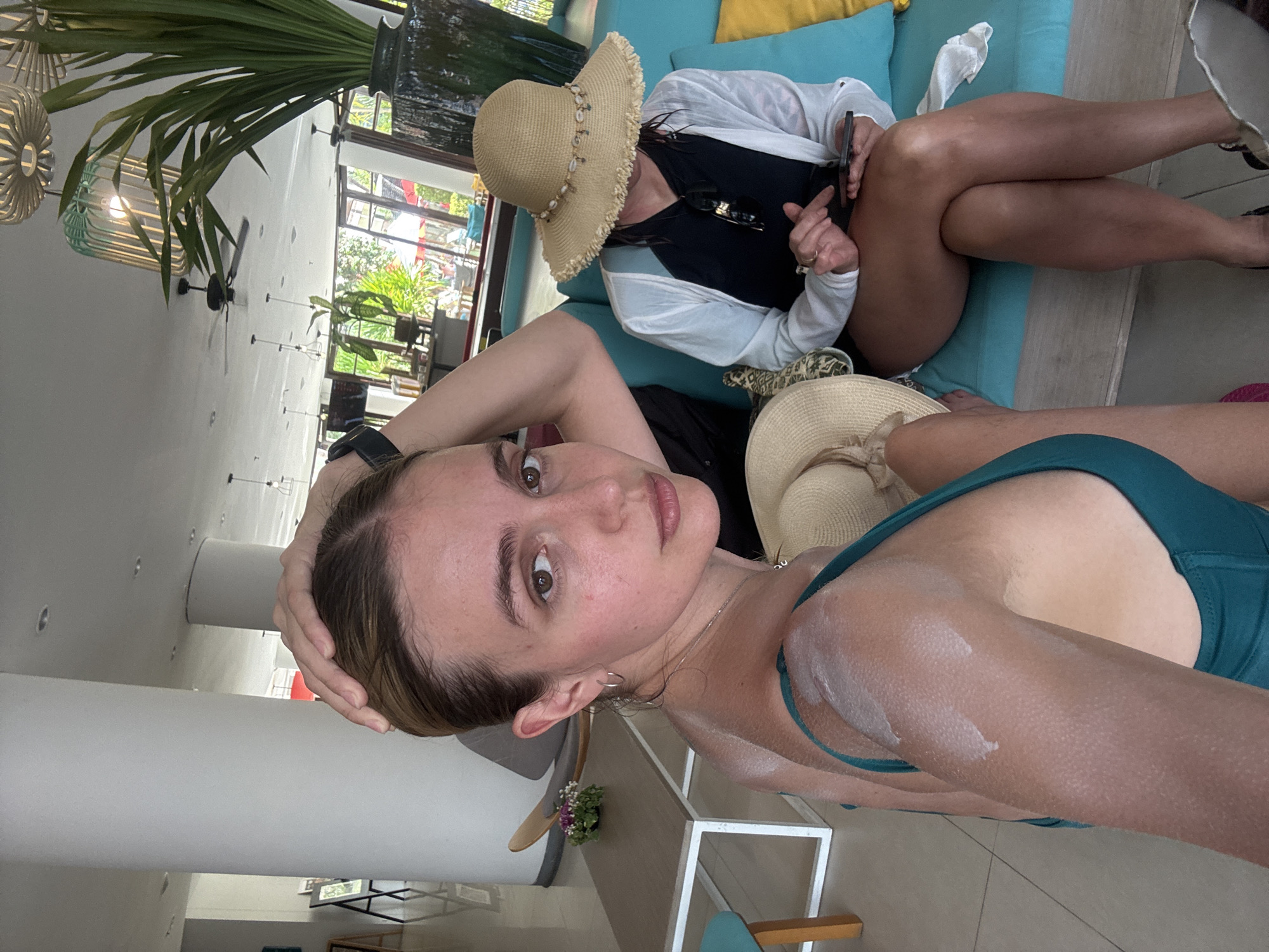 The image shows two women in what appears to be a resort setting. The woman in the foreground is closer to the camera, wearing a green swimsuit and sporting visible sunscreen on her shoulder. She is looking directly at the camera with a slight smile, her hand touching her head. The background woman is sitting on a light blue couch with yellow pillows. She is wearing a black top, white cardigan, and a straw hat adorned with seashells. She holds a dark object, possibly a phone, in her hand.
The location appears to be an indoor/outdoor area with a view of palm trees and other foliage visible through glass windows. A large vase with tropical plants sits near the couch. A table and chairs are partially visible, along with other decorative elements.
Based on the details and the implied resort setting, it is plausible that this image was taken in a location such as a resort or upscale hotel in the city of Patong, Thailand. The specific details of the environment like the blue sofa and the decor, along with the presence of palm trees in the view, are consistent with such a location.