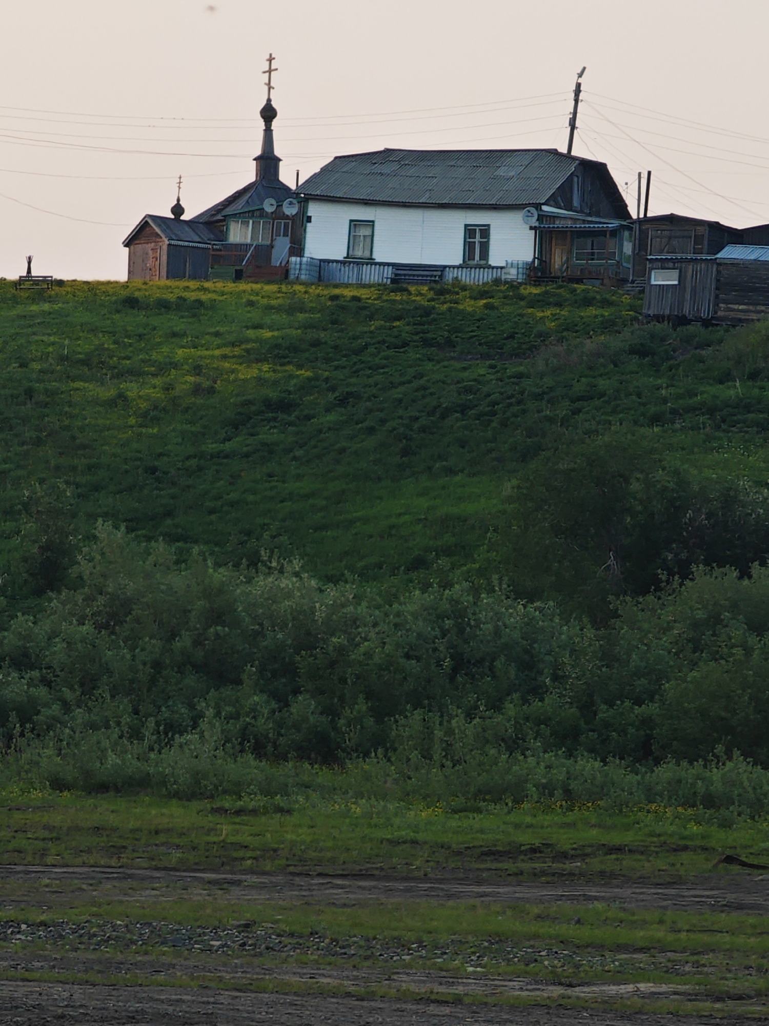 The image depicts a rural scene in Potapovo, Russia, featuring a cluster of buildings on a grassy hill. A prominent white house with a dark, corrugated roof stands centrally. To its left is a dark-colored building with a similar roof and a tall, dark spire topped with a cross, indicative of a church or chapel. Adjacent to this structure is a smaller, wooden outbuilding. Several satellite dishes are visible on the main house and the church building. The hill is covered in lush green grass with scattered patches of yellow wildflowers. In the foreground, a dark, muddy area with some rocks and sparse vegetation suggests a pathway or unpaved ground. The sky is a pale, hazy orange, indicating either dawn or dusk. There are no people or animals clearly visible in the image. Power lines are strung across the upper portion of the scene, supported by wooden poles.