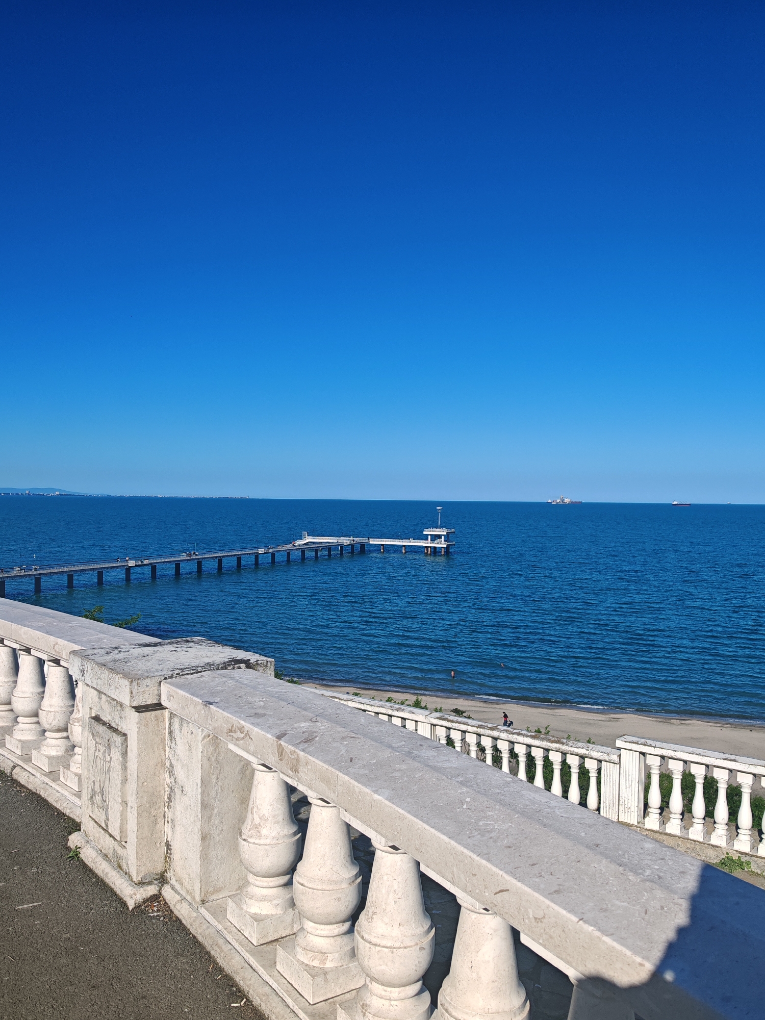 The image captures a scene in Burgas, Bulgaria, overlooking the Black Sea. A concrete balustrade with classical design elements runs along the bottom of the frame. Beyond the balustrade is a sandy beach with a few people visible, including one individual standing near the water's edge. A long pier extends into the sea, culminating in a structure with a platform supported by columns. In the distance, several ships are visible on the horizon. The sky is clear and blue. The overall environment suggests a sunny day at a coastal location.