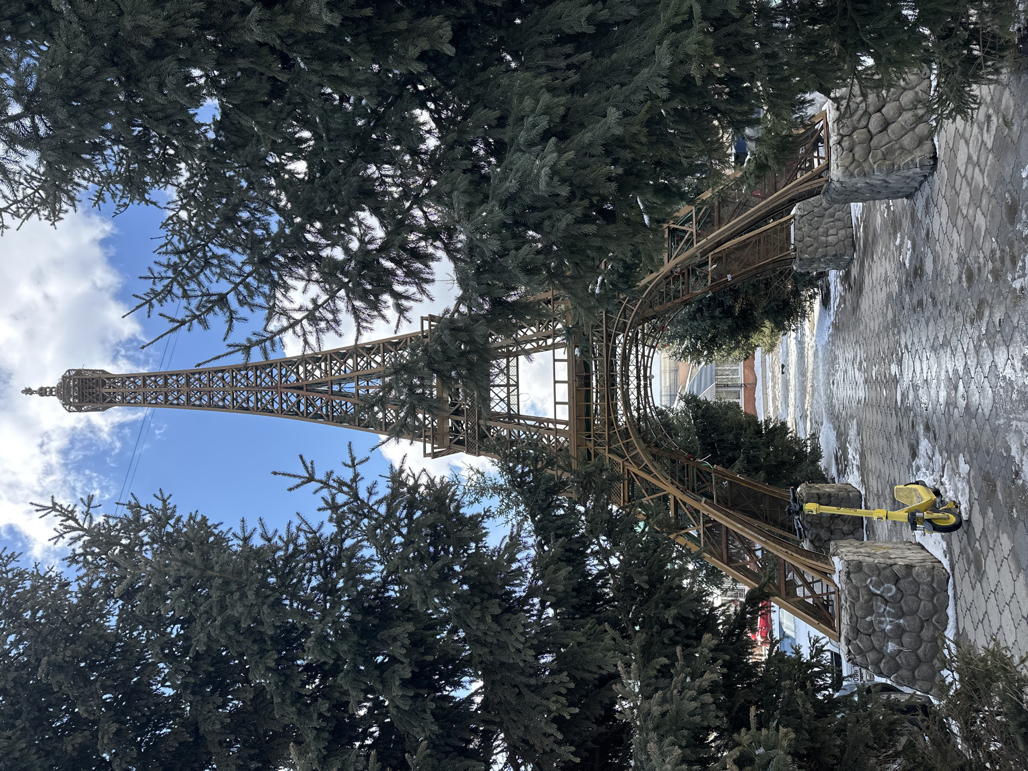 The image depicts a model Eiffel Tower within an archway structure, positioned in an outdoor setting likely in Krasnoyarsk, Russia. The tower is constructed from a brown, possibly metal, framework, and it is partially obscured by evergreen trees in the foreground. The archway appears to be of similar material to the tower, supported by stone-like pillars. A paved walkway extends away from the archway, showing remnants of snow or ice. A yellow scooter leans against one of the stone pillars near the archway. The sky is visible through gaps in the trees and the tower's structure, showing a partly cloudy sky.