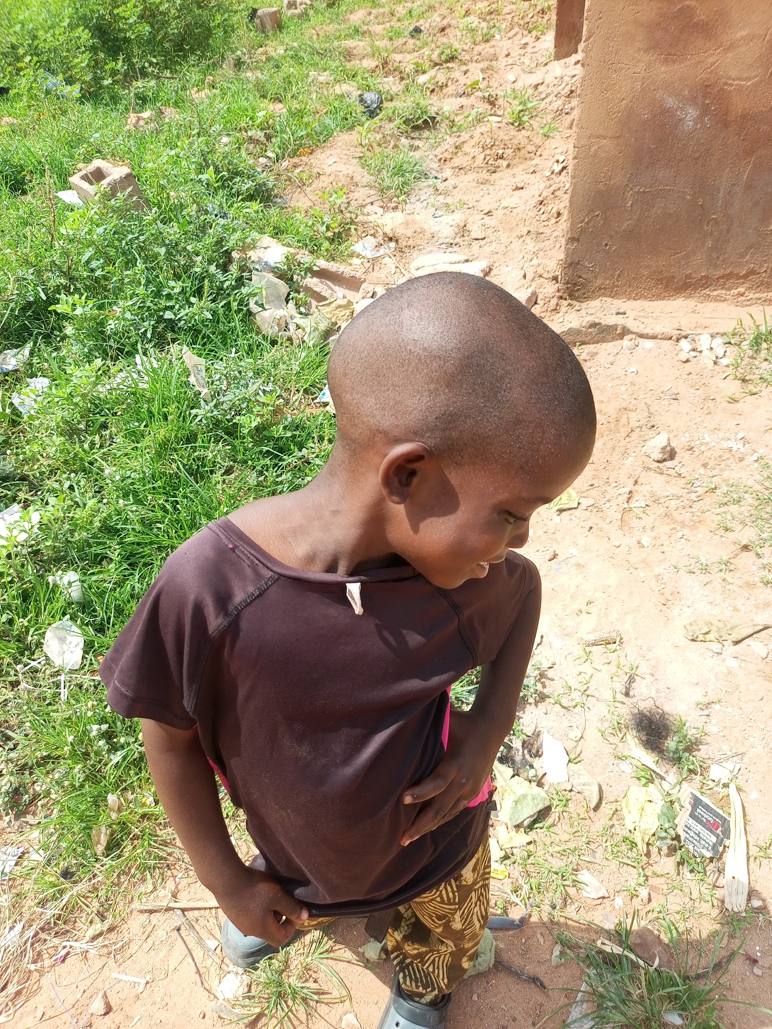 A child with a shaved head and dark skin, wearing a dark brown t-shirt and patterned trousers, is standing outdoors on a dirt ground with sparse green vegetation. The child is looking downwards and to the right, with their right hand resting on their hip. In the background, a rough-textured, light brown wall of a building is visible, and to the left, there is an area with denser green grass and some scattered debris, including what appears to be bricks and plastic. The overall setting suggests an environment in Gombe, Nigeria, possibly a residential area or a less developed part of the city, characterized by its unpaved ground and the presence of construction materials and natural vegetation.