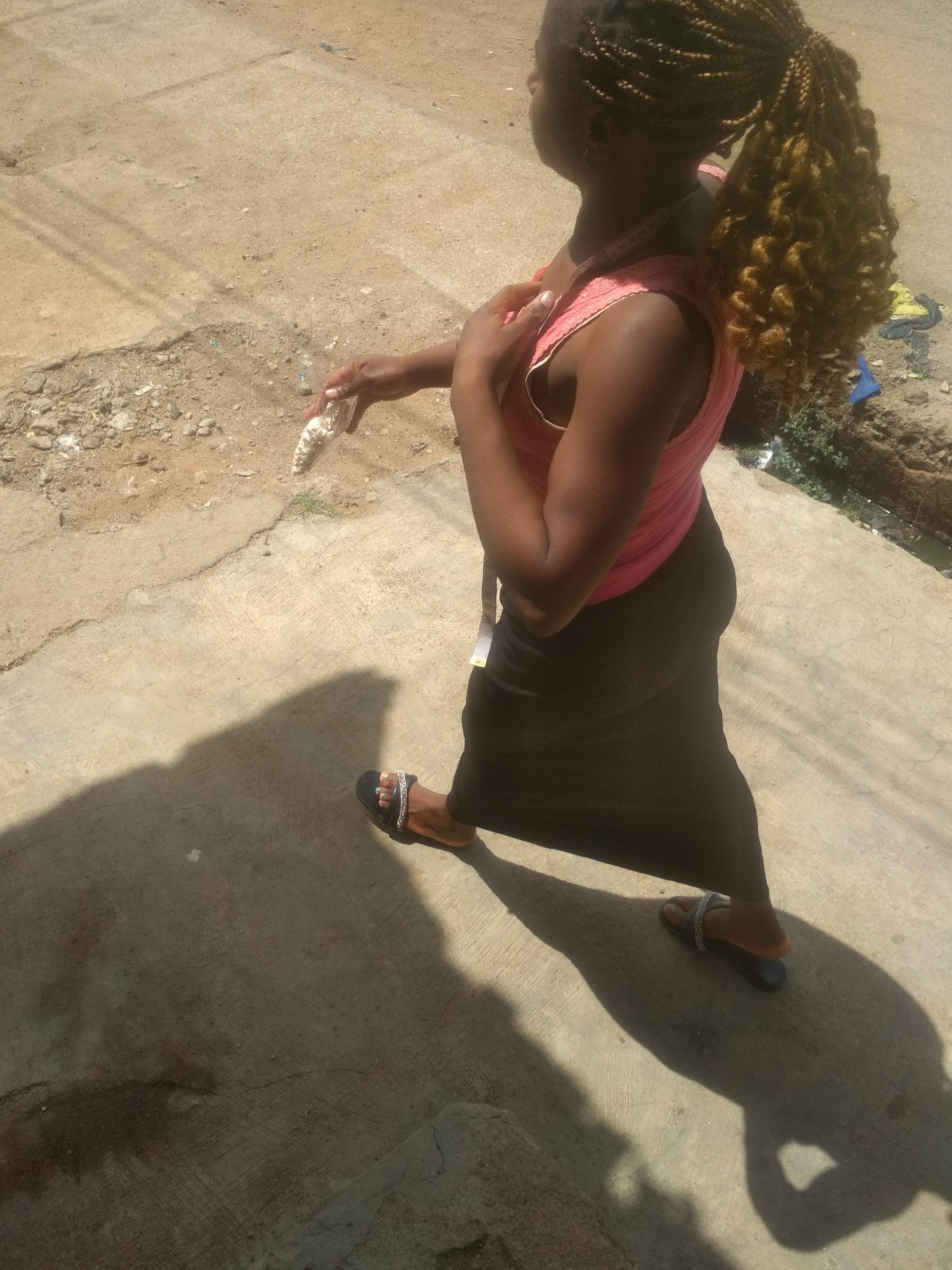The image shows a woman standing in an outdoor location in Jos, Nigeria. She has dark skin, with her hair styled in braided pigtails gathered into a curly ponytail. She is wearing a pink sleeveless shirt and a dark-colored long skirt. A measuring tape hangs around her neck. In her right hand, she holds a small plastic bag. She is standing on a concrete surface with cracks and patches of dirt, likely a sidewalk or paved area. Her shadow is cast on the ground. In the background, there is a suggestion of vegetation and some scattered debris. The image is taken in daylight, casting strong shadows.