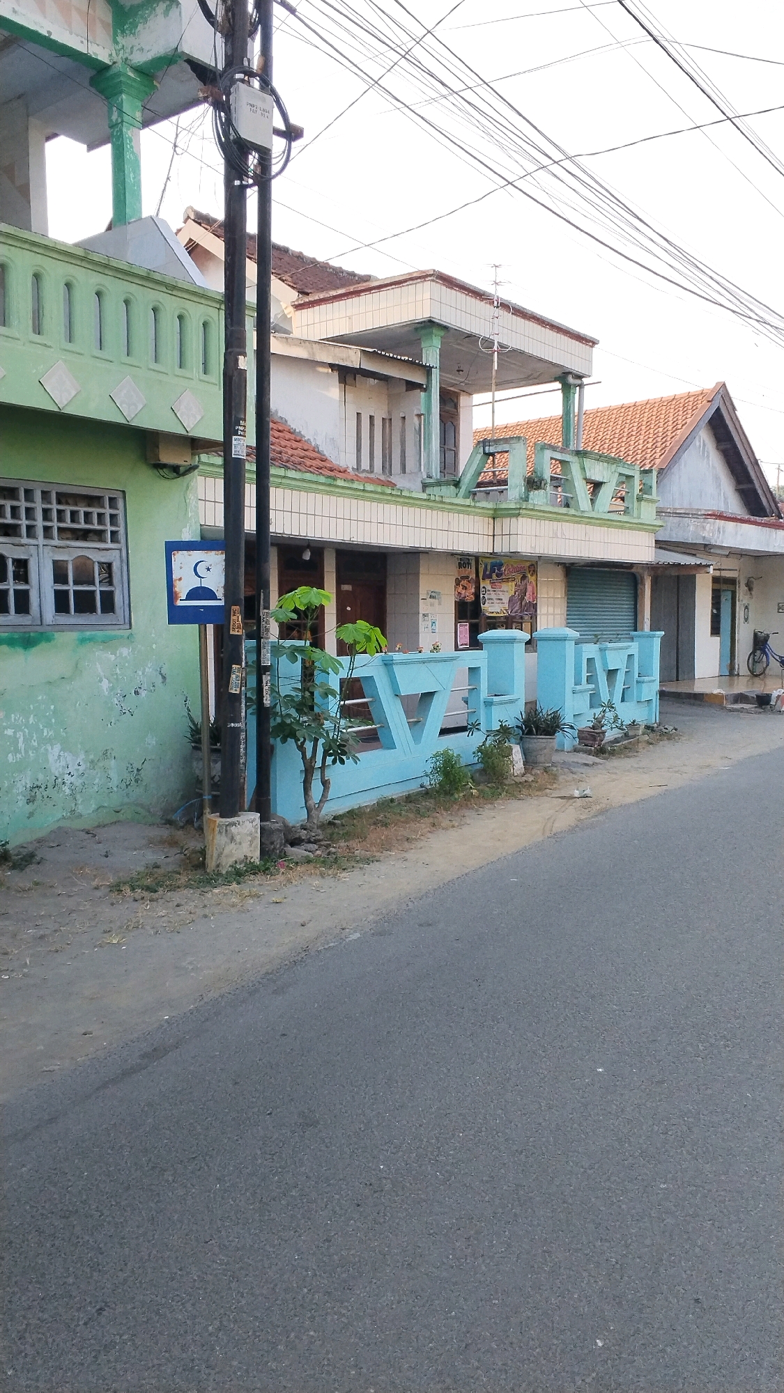 The image depicts a street view in Penompo, Indonesia, showing the front of a building with a light green facade. A black utility pole stands in front of the building, covered in electrical wires. A blue sign with a crescent moon and star symbol is attached to the pole. Adjacent to the building is a light blue decorative fence. The building appears to have multiple stories and a tiled roof. In the background, there are more buildings and electrical wires crisscrossing the sky. The ground in front of the building is a dirt and gravel path, leading to an asphalt road. The scene appears to be in an urban or semi-urban environment, with the sun casting a warm light.